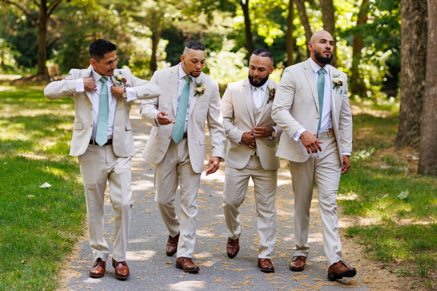 Four groomsmen in tan suits with mint ties walking confidently down a tree-lined path outdoors.