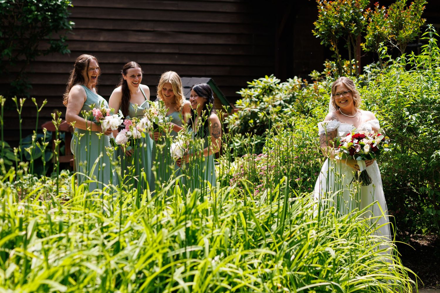 Bride in white gown with bridesmaids in sage green dresses holding bouquets in a lush garden setting.