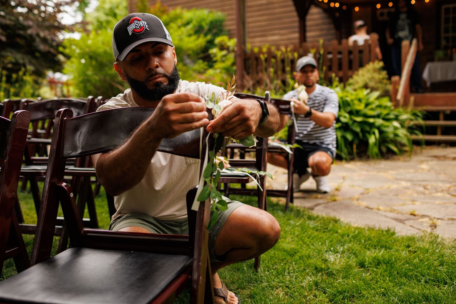Man in Ohio State cap arranges chairs outdoors at a garden venue with string lights and lush greenery.