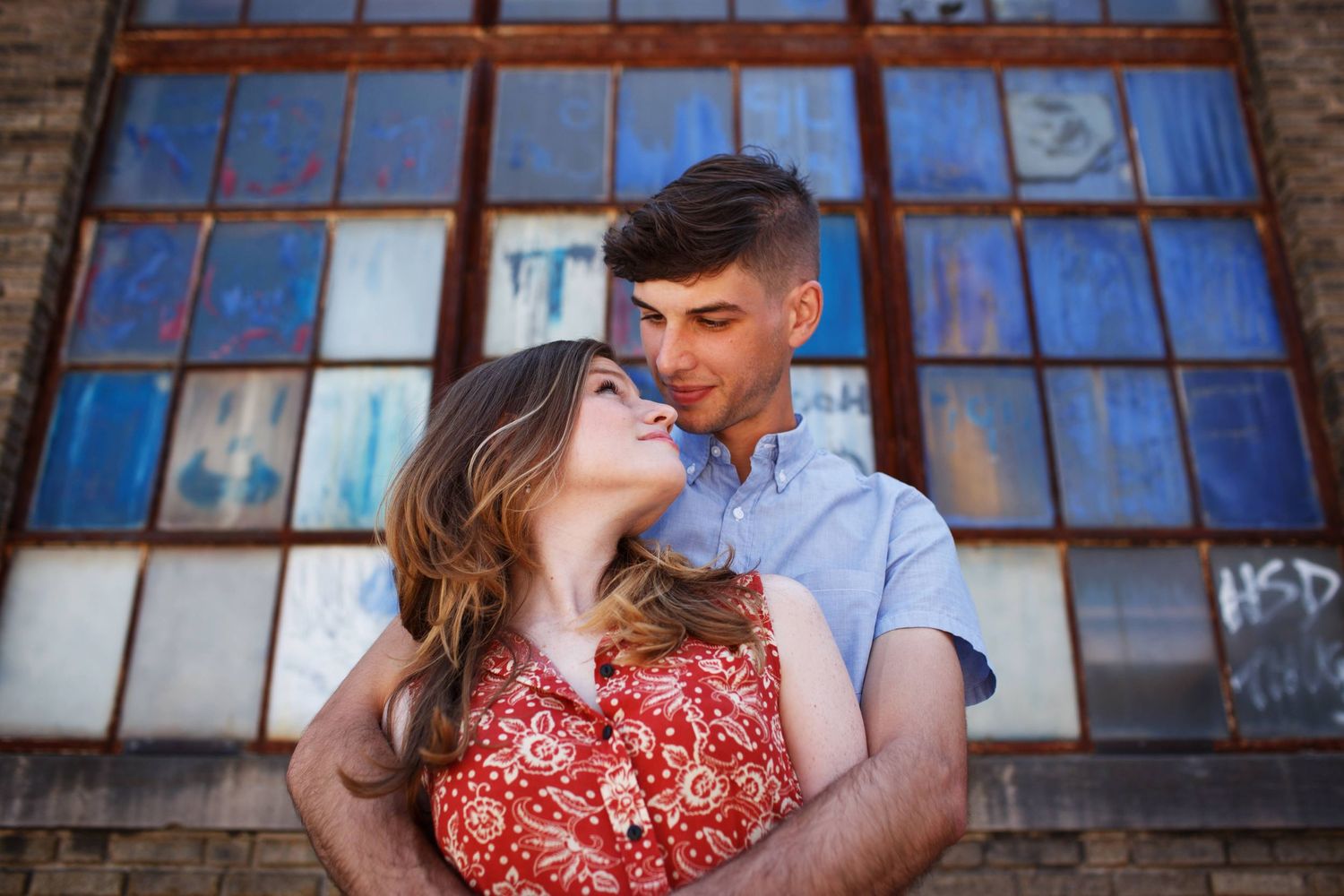 A couple shares a tender moment in front of an industrial window with blue panes and brick walls.