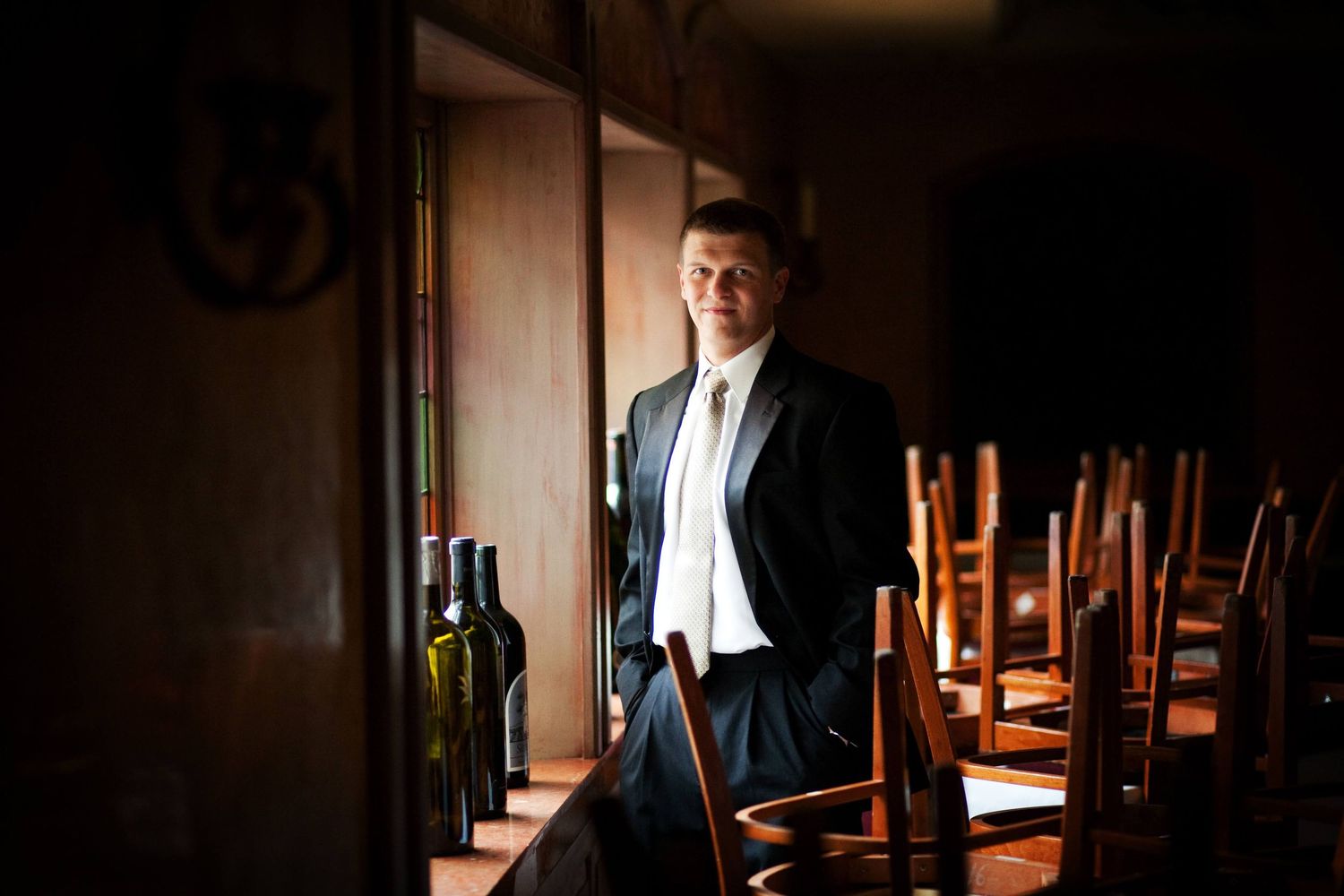 A figure in a formal suit stands in a dark restaurant dining room with stacked chairs and wine bottles on display.