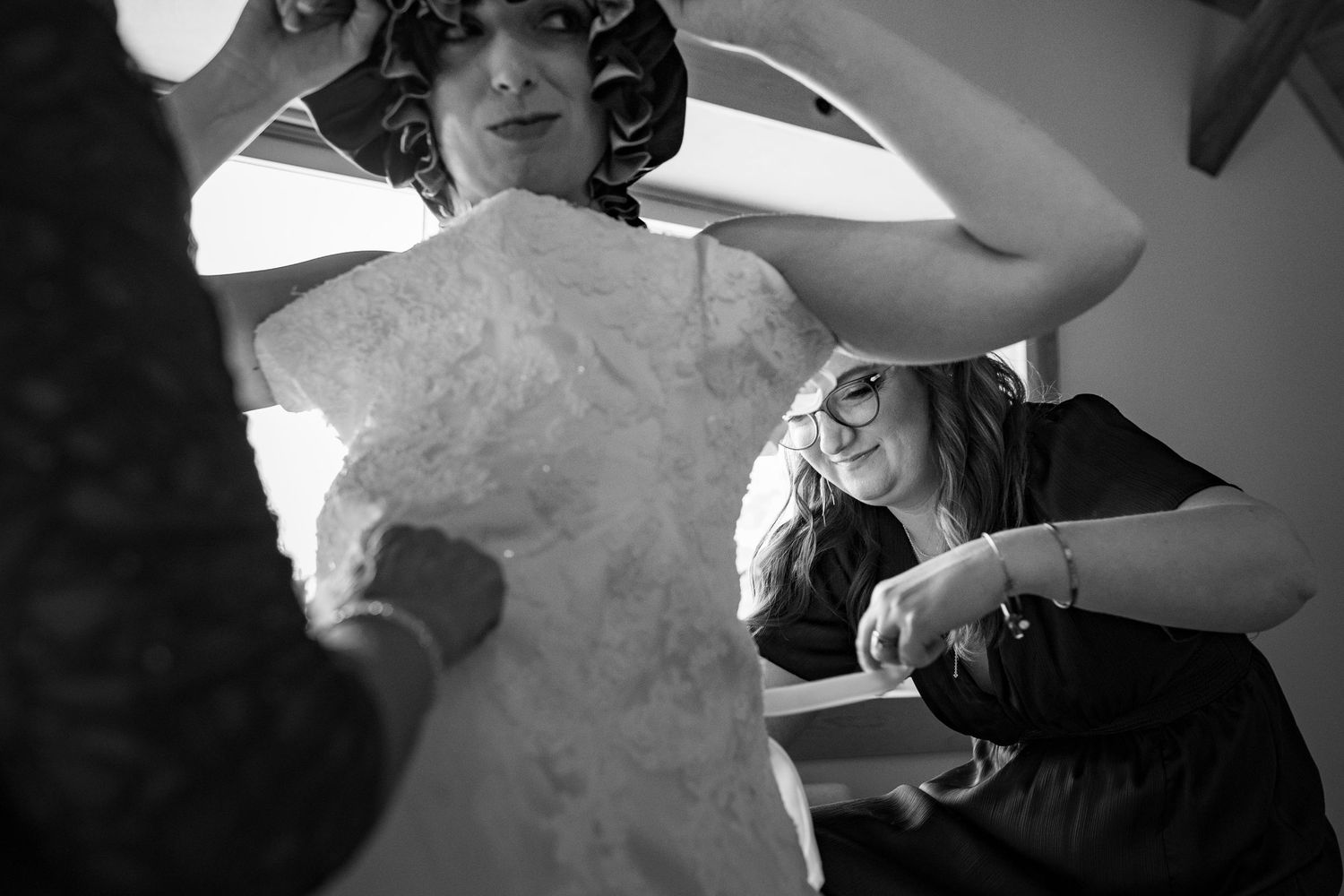 Black and white photo of bride being helped into wedding dress by two women during bridal preparations.