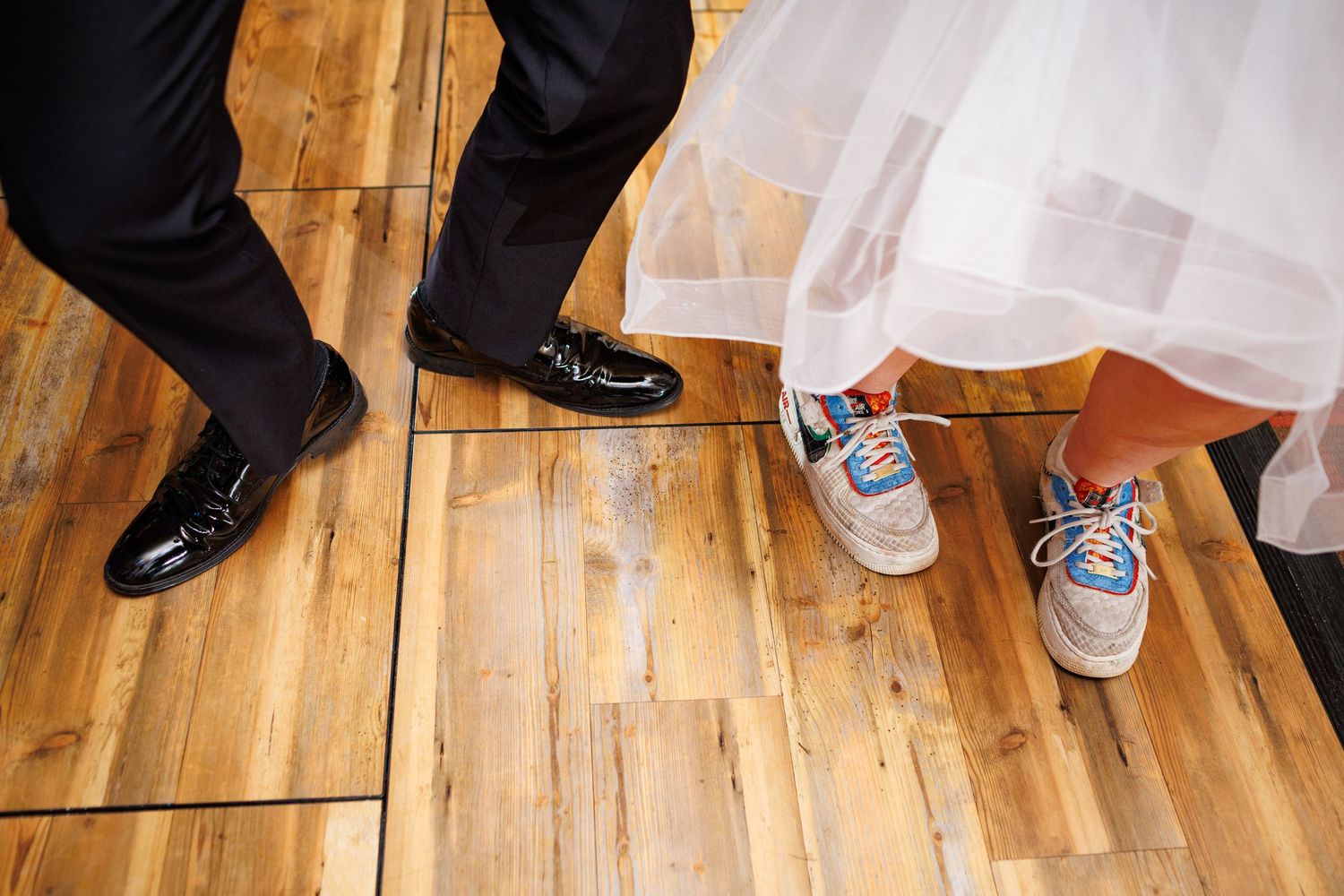 Close-up of bride wearing colorful sneakers and groom in black dress shoes on wooden dance floor.