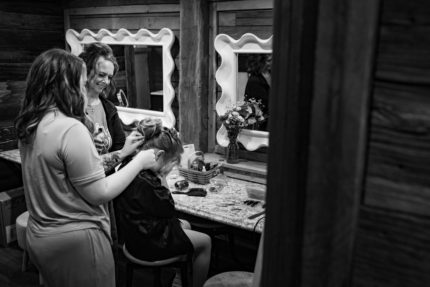 Black and white photo of a hairstylist styling a seated woman's hair in a rustic vanity room with mirror.