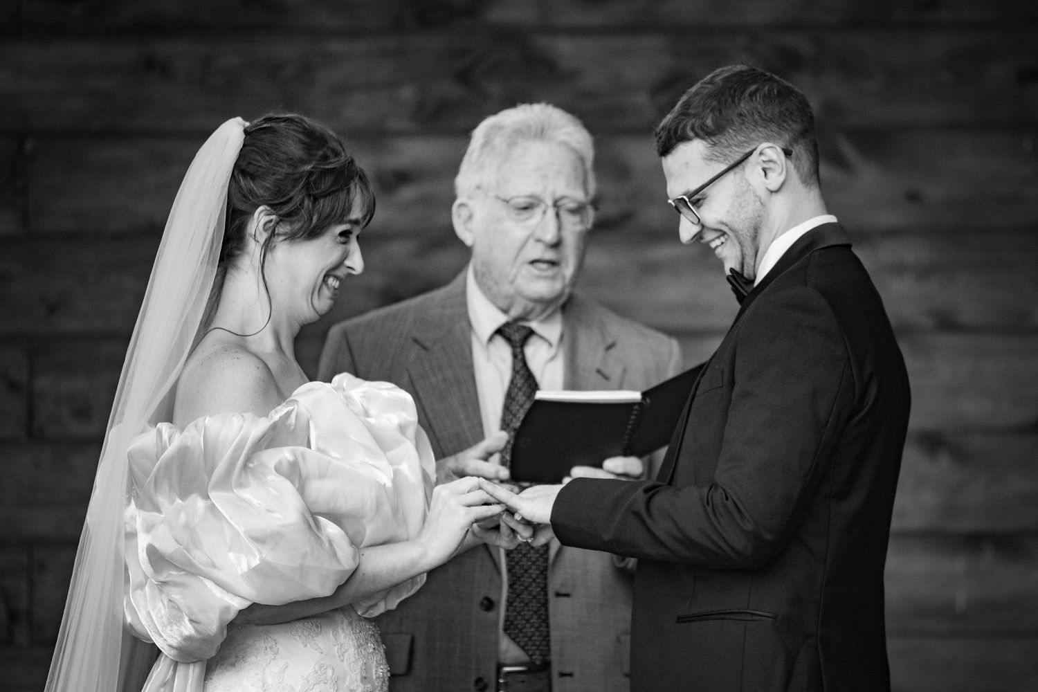 Black and white wedding ceremony photo of bride and groom exchanging rings before an officiant.