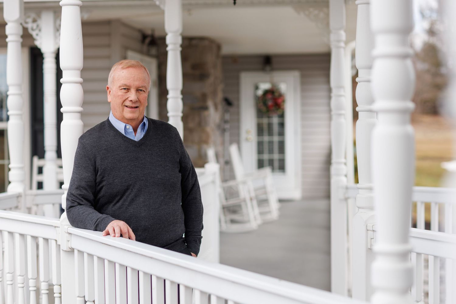 Someone stands on a white wraparound porch with rocking chairs and decorative columns on a traditional house.