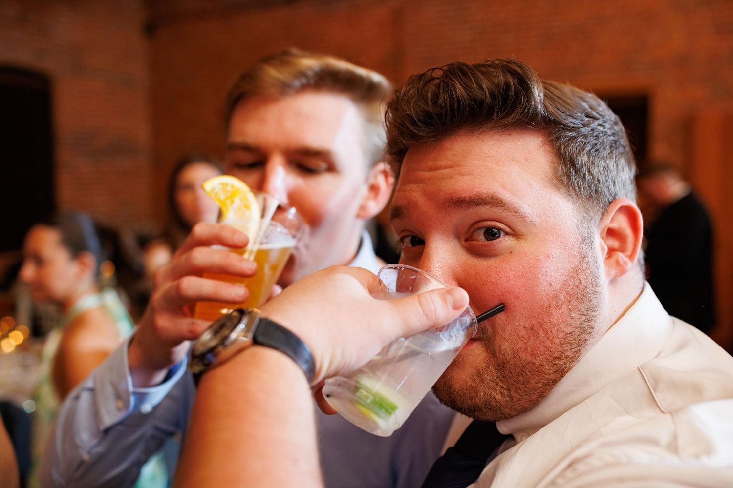 Wedding guest humorously drinks from glass at lively reception with guests celebrating in background.