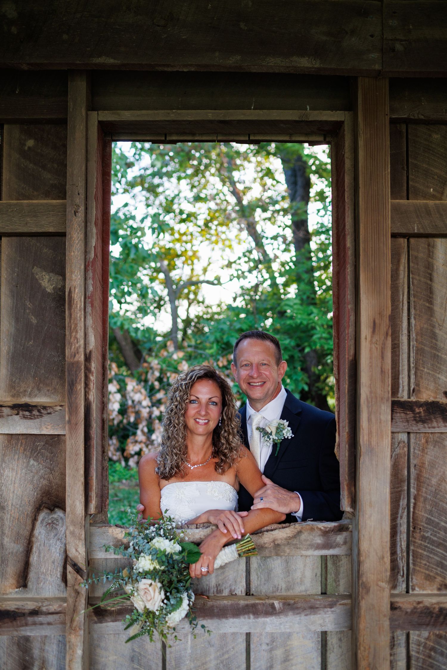 Smiling bride and groom pose together in a rustic wooden barn window on their wedding day.