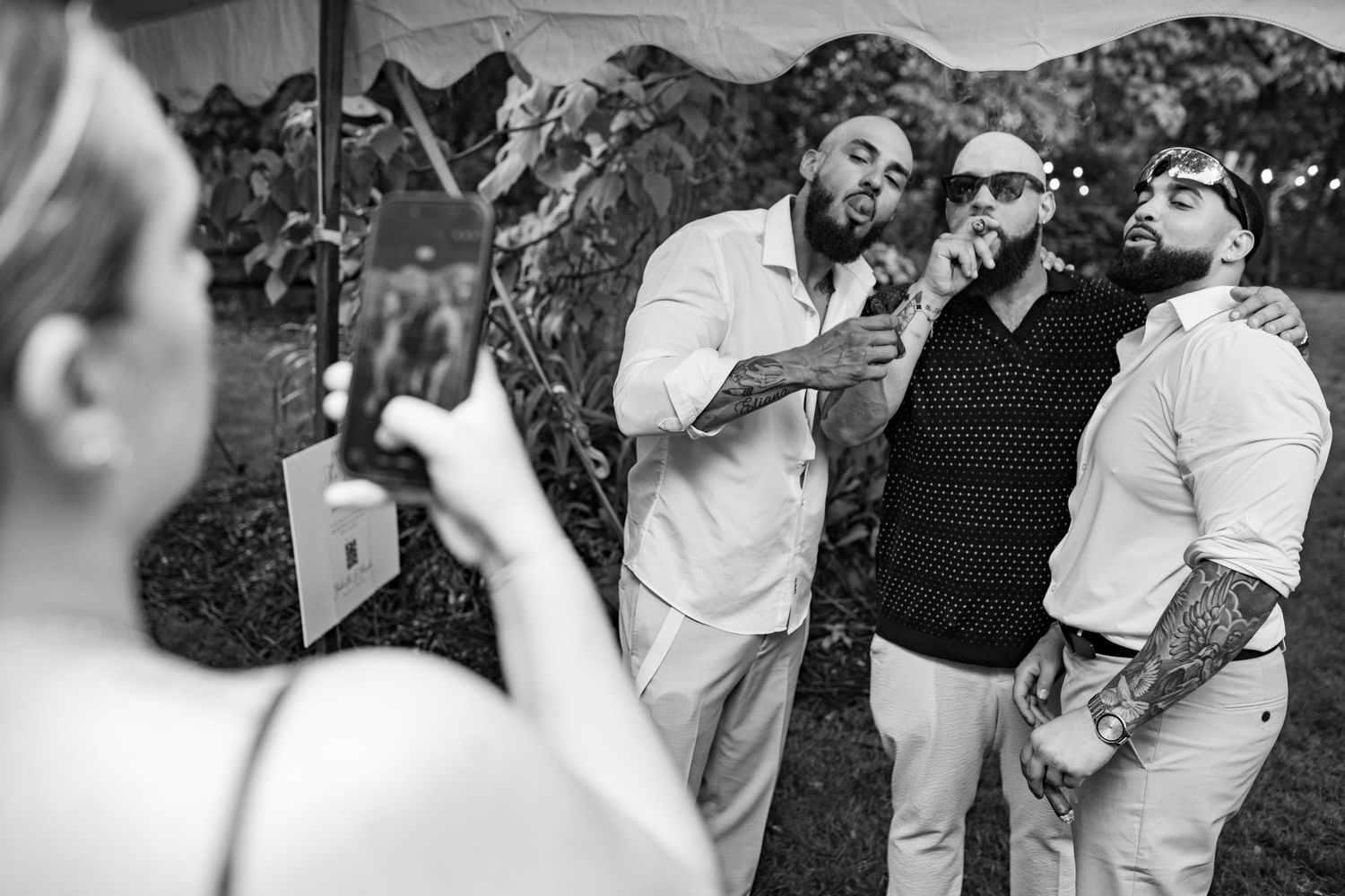 Black and white photo of three men posing with cigars while a woman photographs them at a wedding.