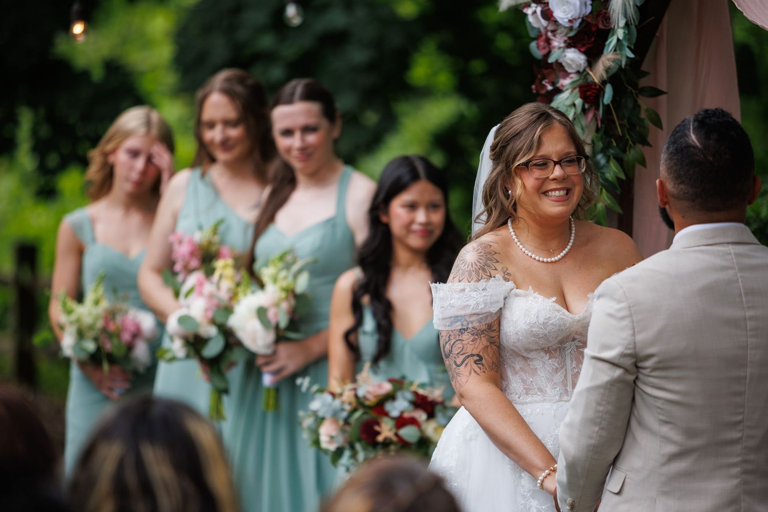 Bride in strapless gown smiles at groom during outdoor wedding ceremony with bridesmaids in mint dresses.