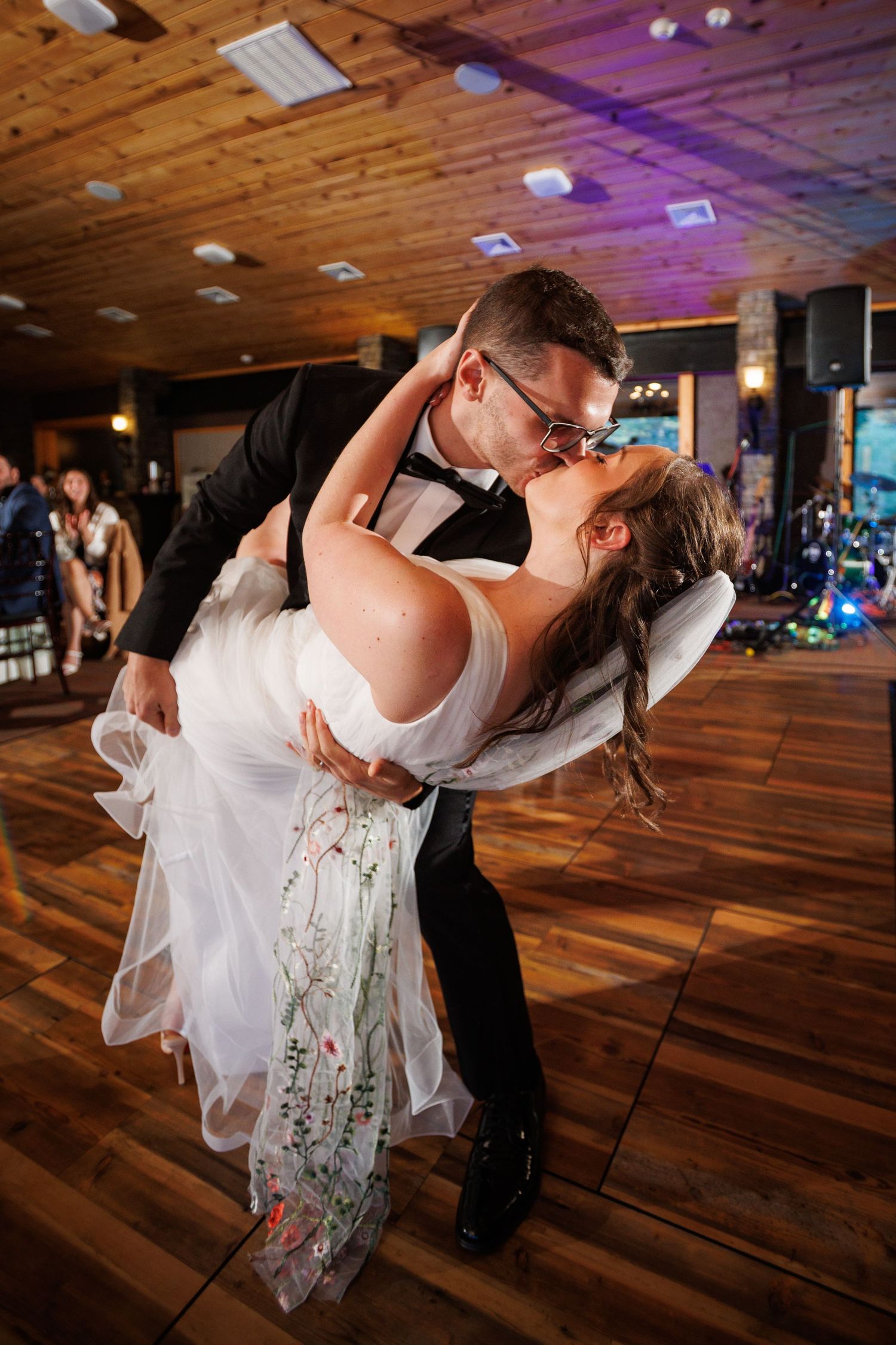 Groom dips bride for romantic kiss on wooden dance floor during first dance at wedding reception.