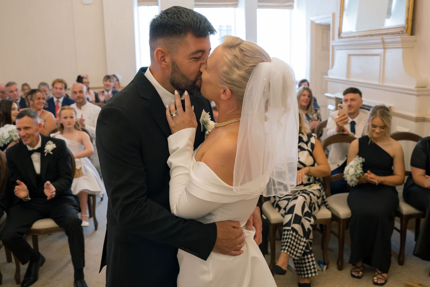 Bride and groom share first kiss at wedding ceremony while guests celebrate in background.
