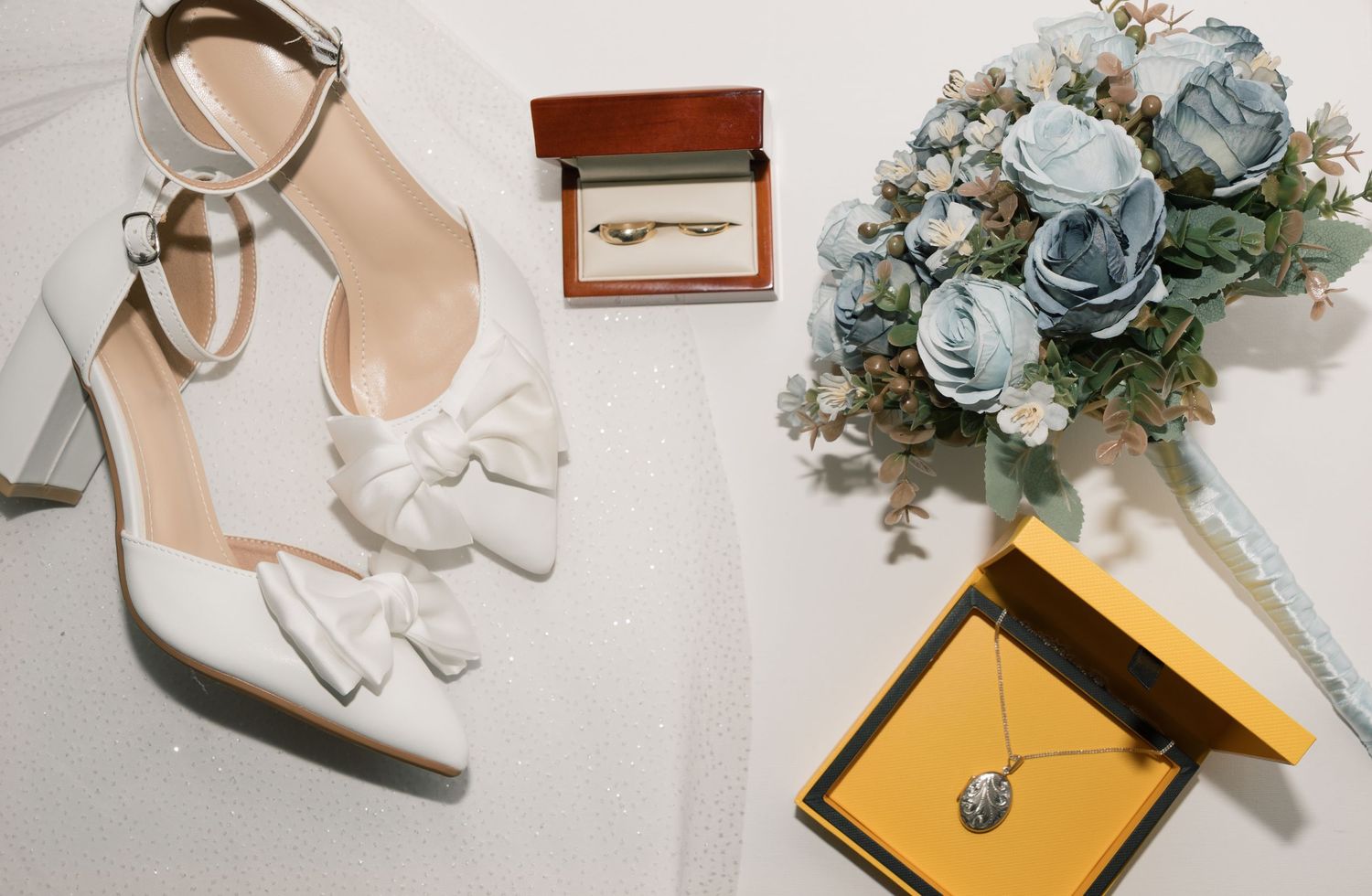 Flat lay of wedding accessories including blue flowers, white shoes, and gold invitation details on a light background.