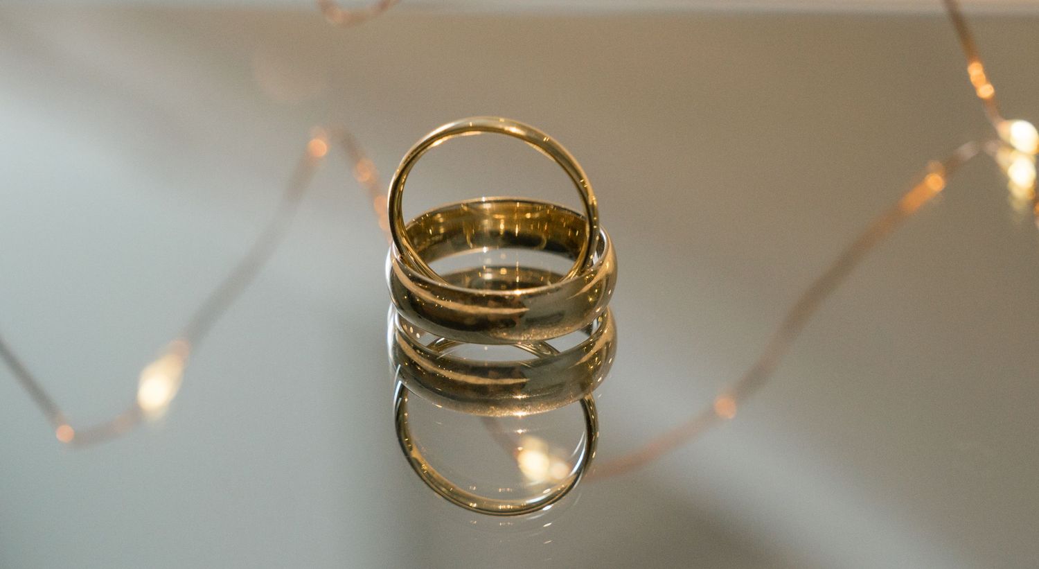 Gold wedding band ring reflecting on glossy surface with soft bokeh lights in background.