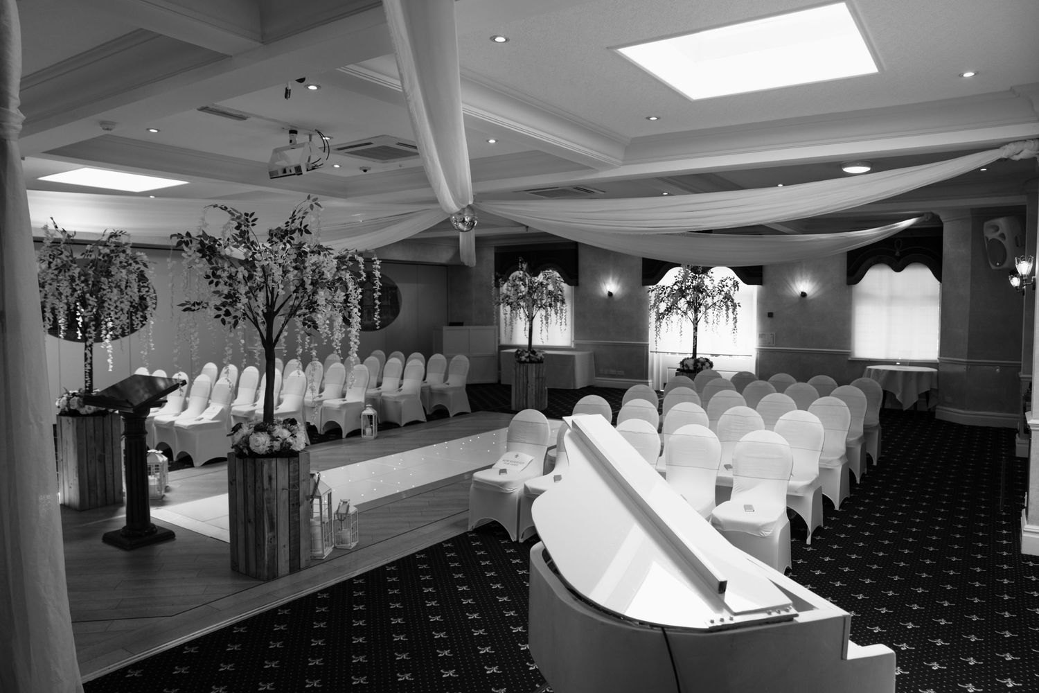 Black and white photo of elegant wedding ceremony room with white chairs and tall tree decorations.