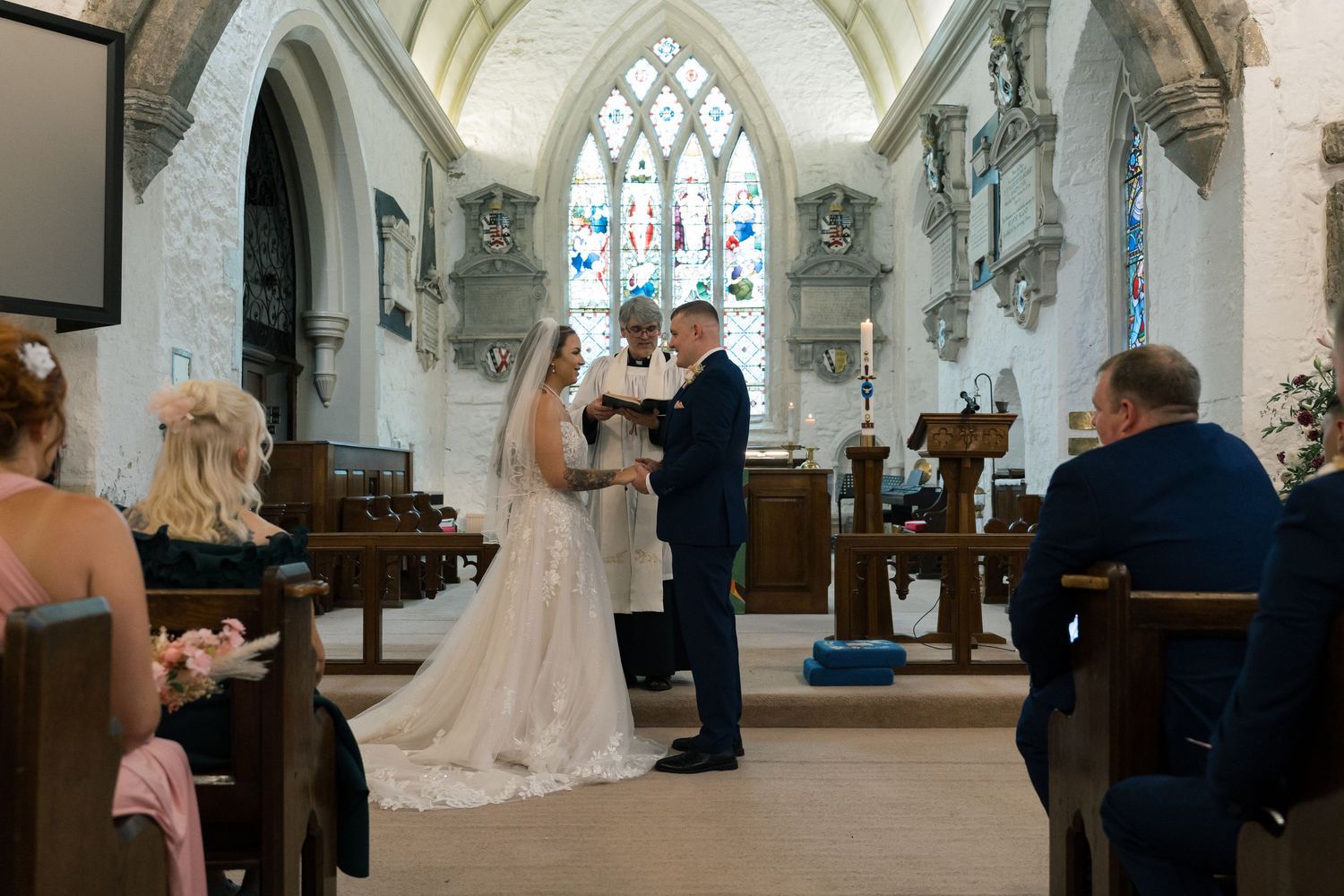 A couple exchanges wedding vows at the altar of a beautiful gothic church with arched stained glass windows and stone walls.