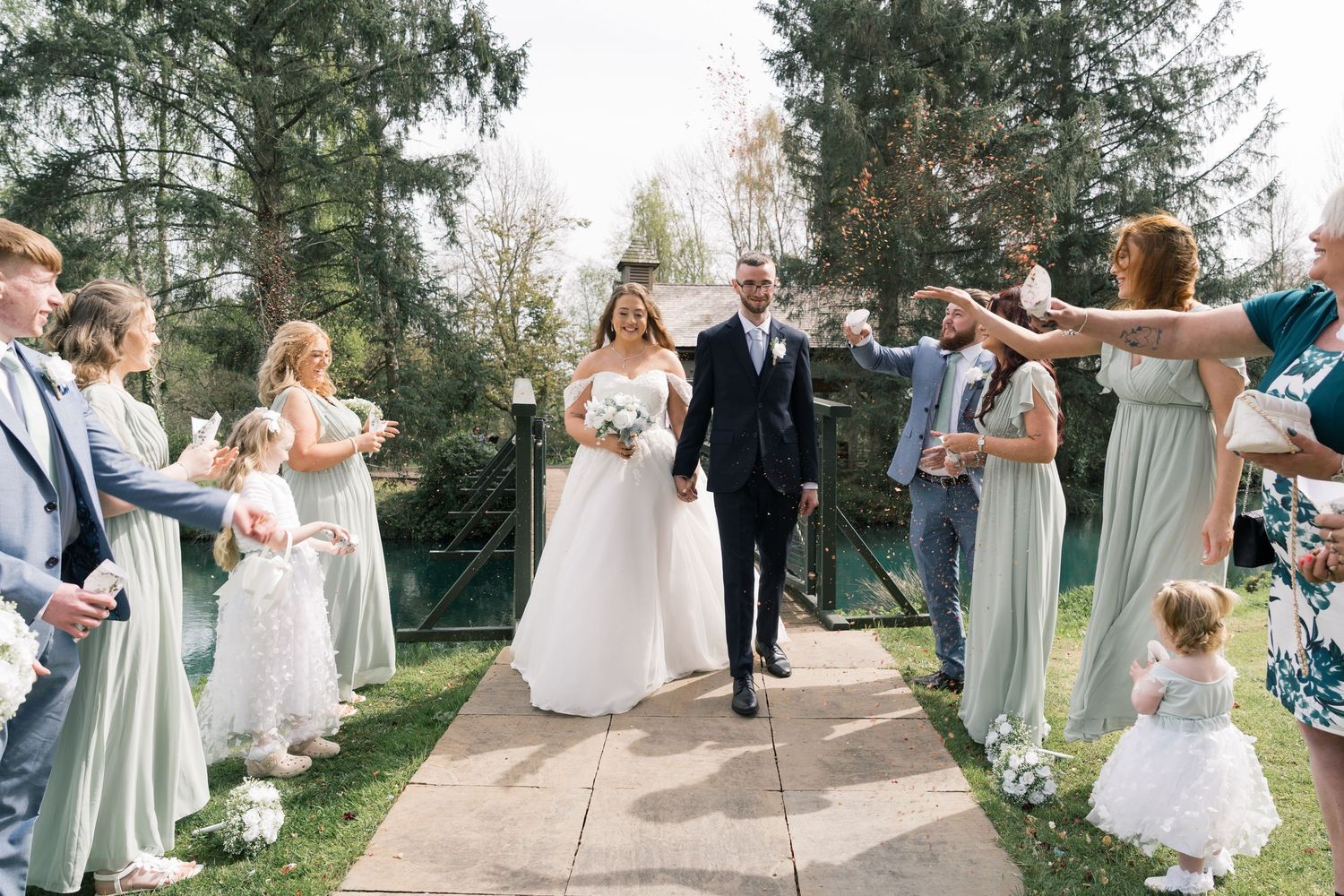 Wedding party in sage green dresses and white gowns walk together on a garden path during an outdoor celebration.