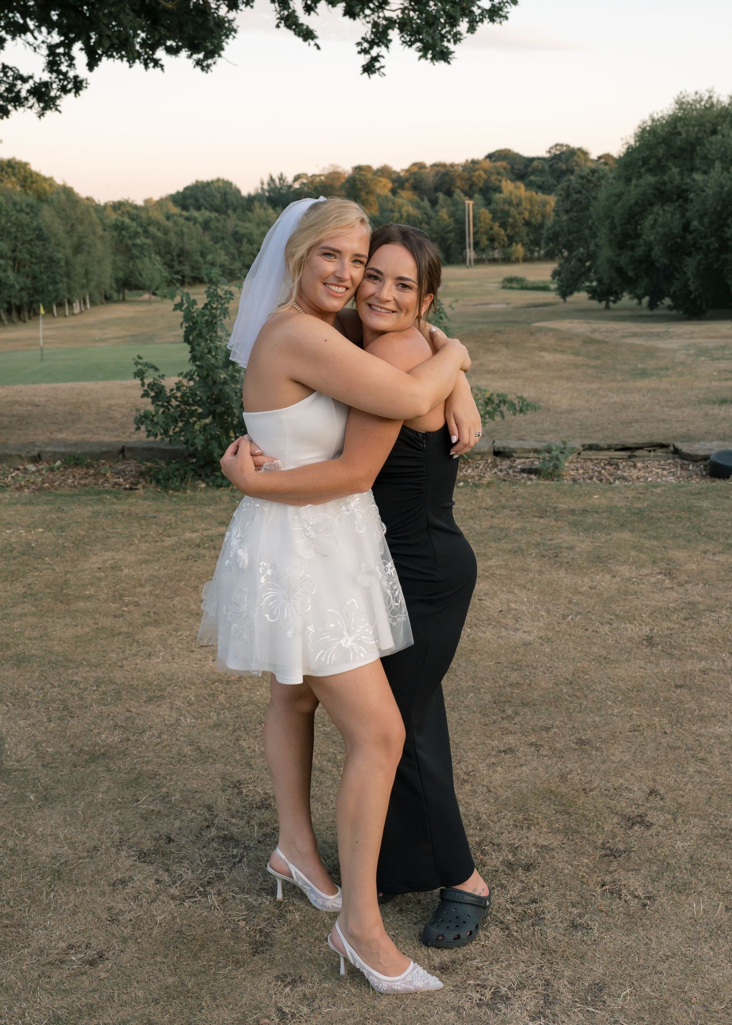 Bride in white mini dress and veil hugging a guest in black gown outdoors at golden hour.