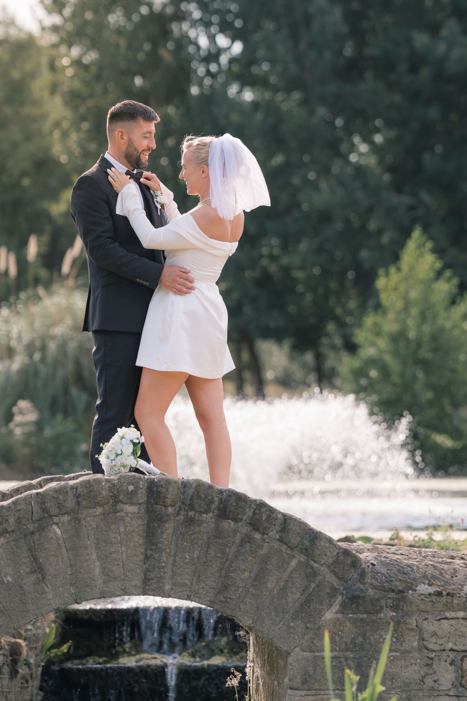 Bride and groom embracing on a stone bridge in a garden, bride wearing a short white dress and veil.