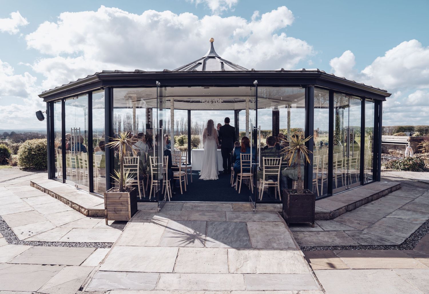 Glass gazebo decorated for an outdoor wedding ceremony with scenic views.