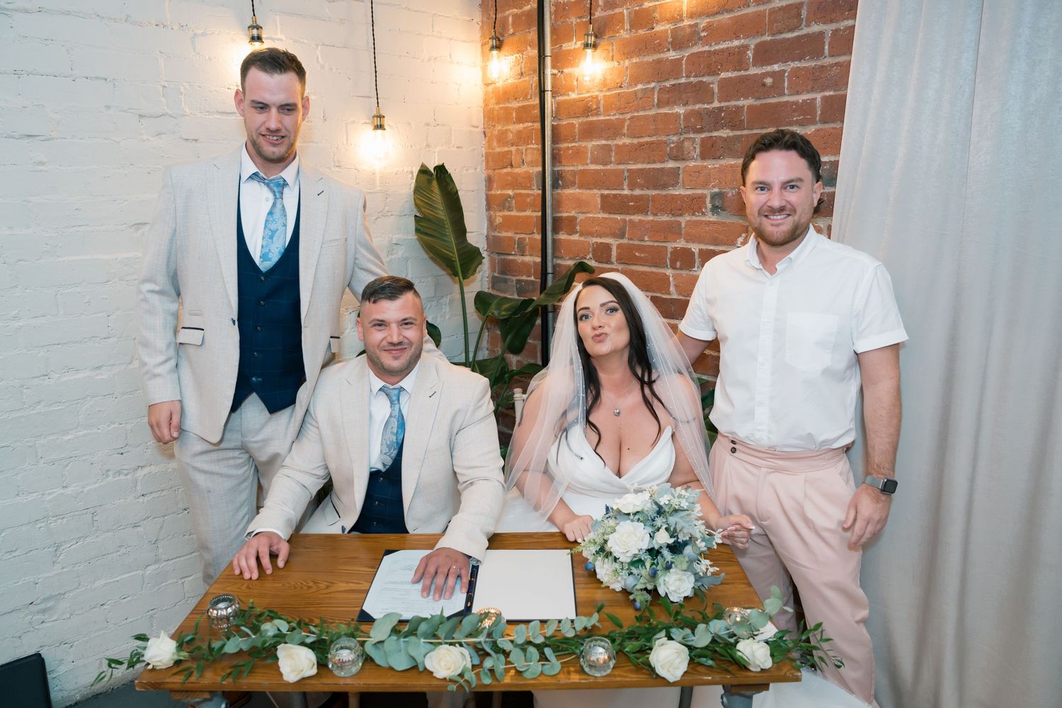 Wedding party gathered around a rustic wooden table with floral decorations and white brick wall backdrop.