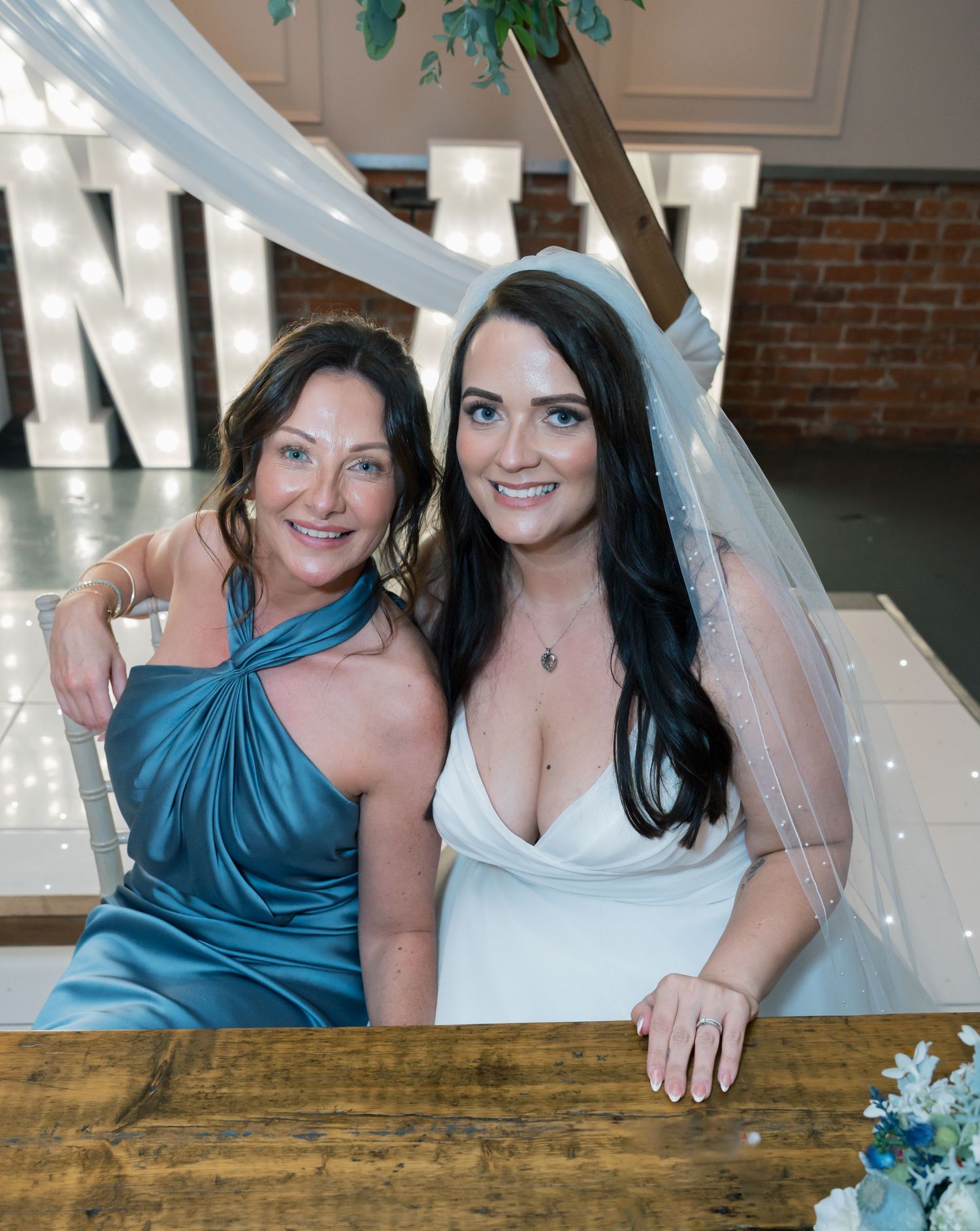Two guests pose together at an event with light blue decor and illuminated letters in the background.