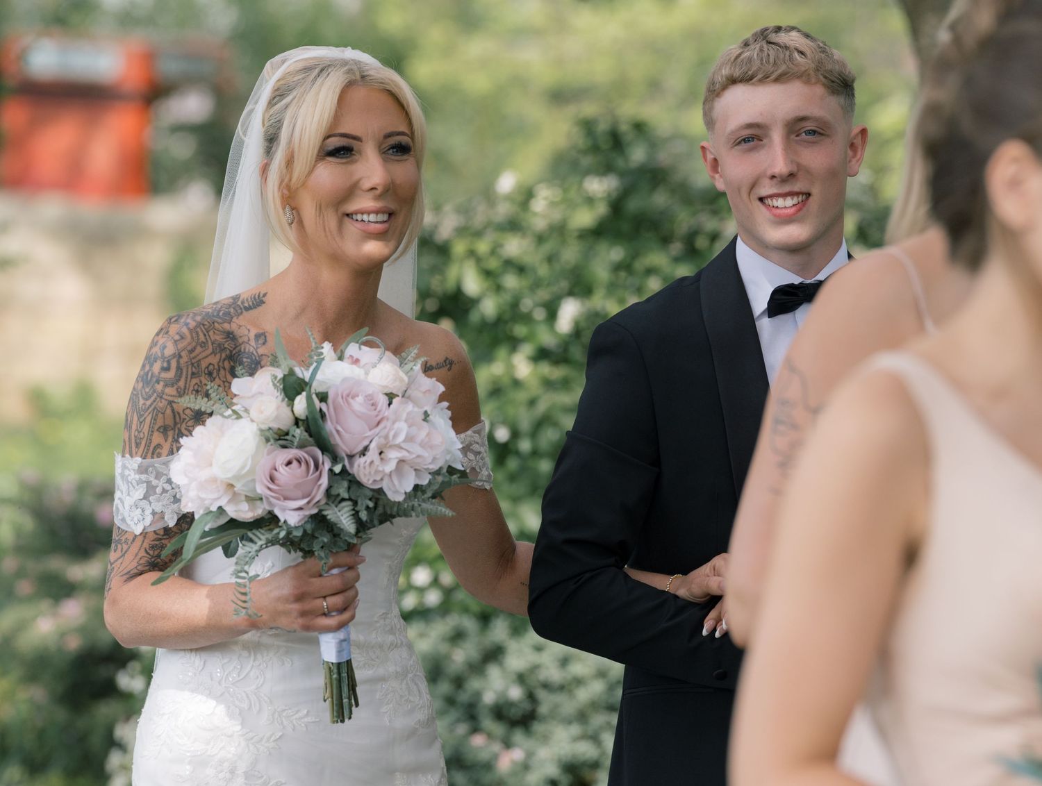 Bride in lace wedding dress holding pink bouquet smiles with groom in black tuxedo during outdoor ceremony.