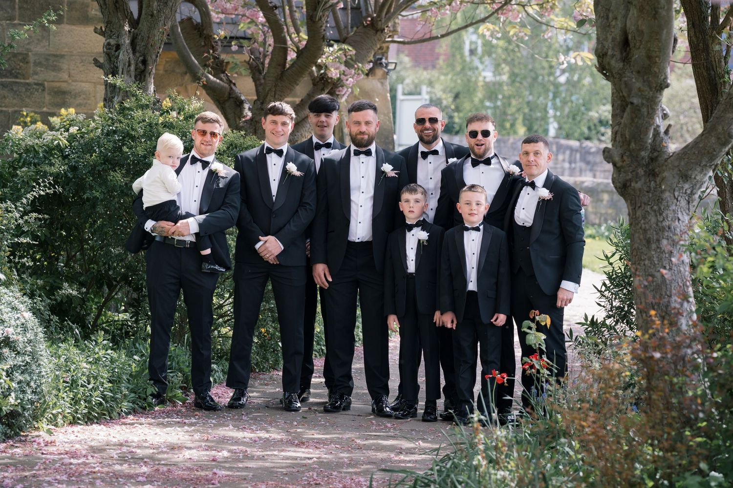 Groomsmen in black tuxedos pose together outdoors in a garden setting for a formal wedding party photo, in Leeds.