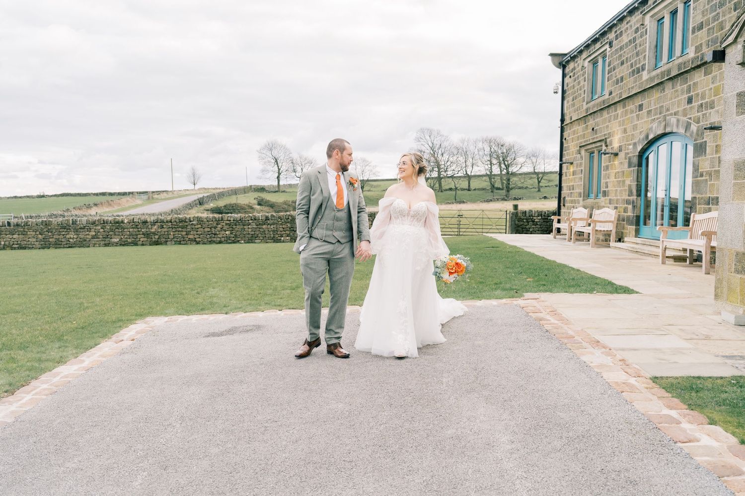 A couple in wedding attire walks together on a path outside a stone building on their special day.