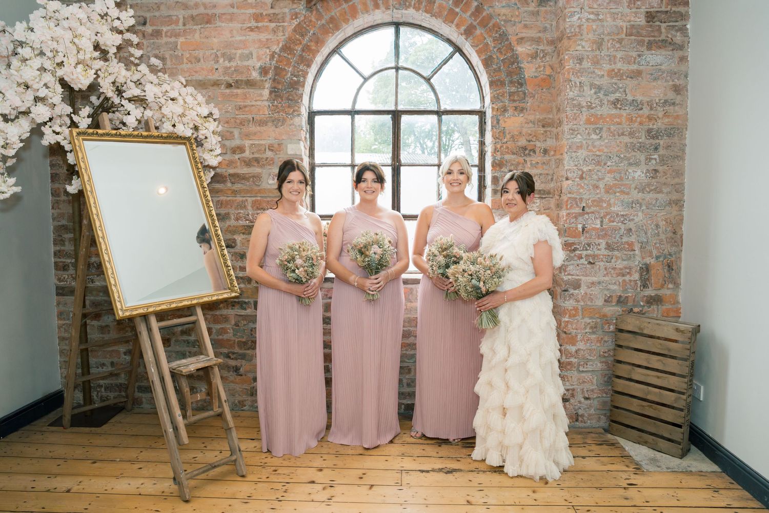Wedding party in pink bridesmaid dresses and ivory gown pose by rustic brick wall with arched window and easel.