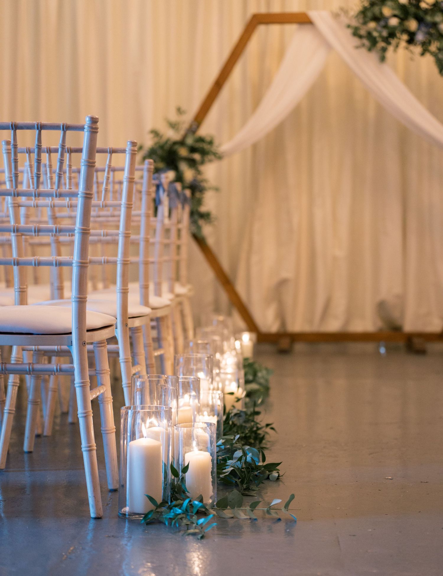 White Chiavari chairs arranged in rows with candles and greenery for an elegant indoor wedding ceremony setup.