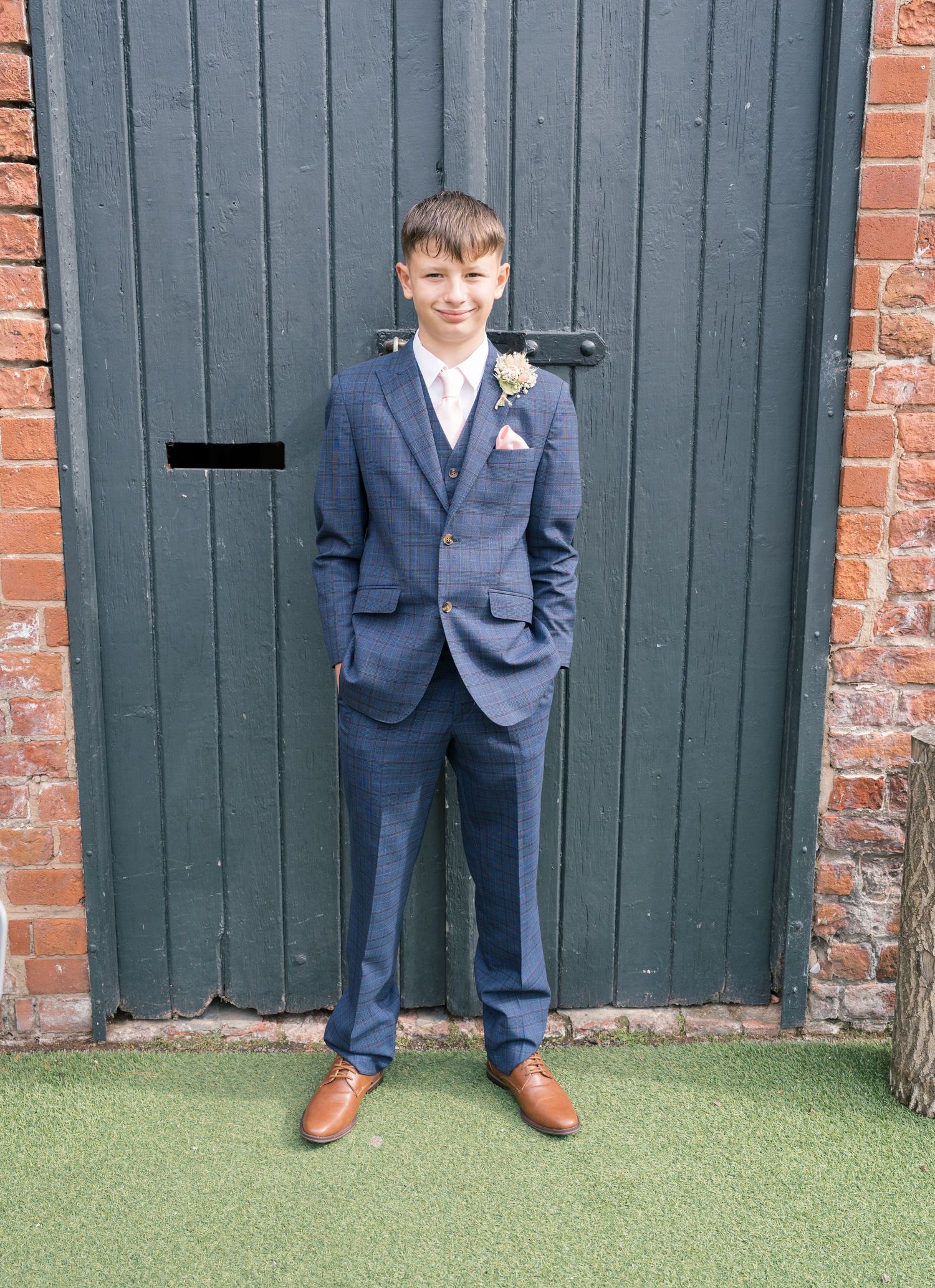 Young attendant in navy suit and brown shoes poses against green door at wedding venue.