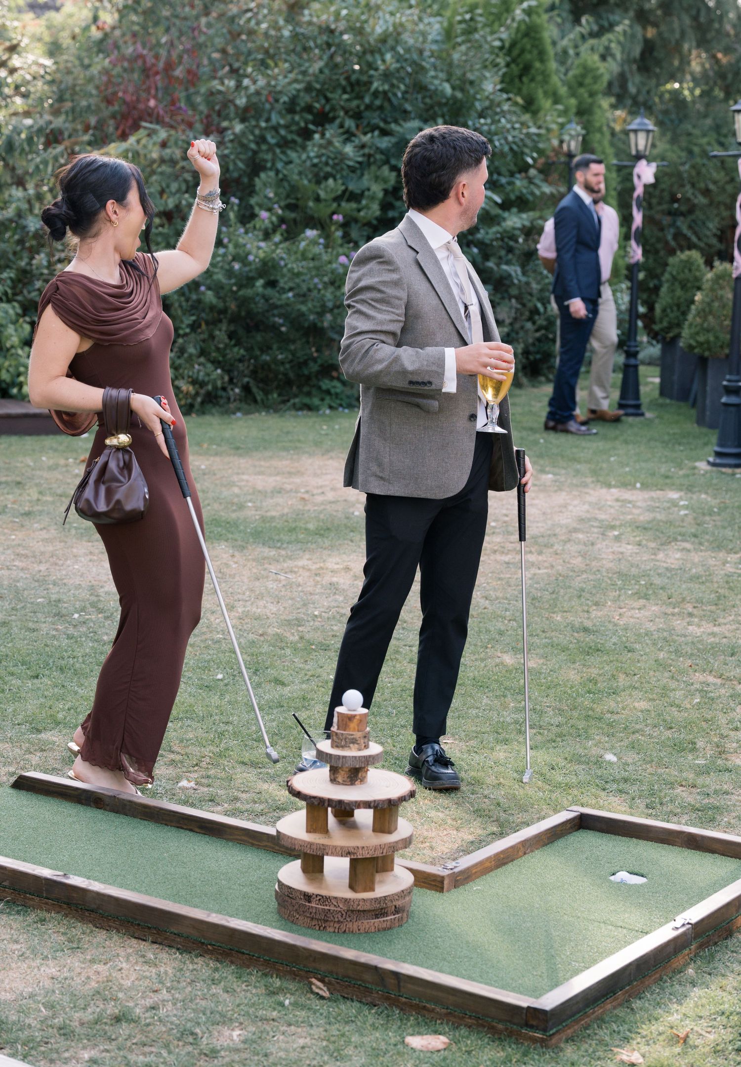 Wedding guests play miniature golf on a portable course set up on grass during outdoor reception.