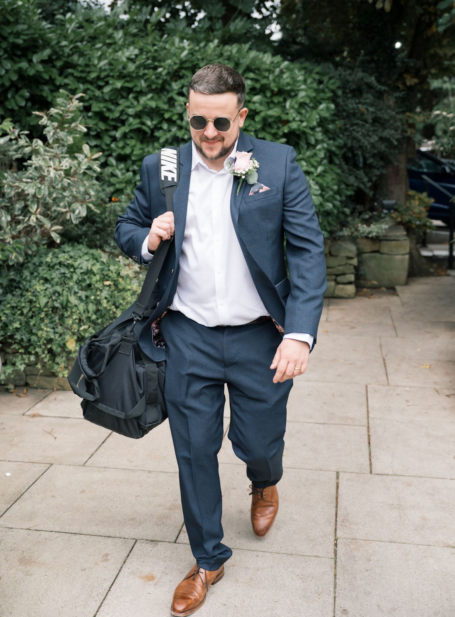 Stylishly dressed guest in navy suit and brown shoes walking with a duffle bag at outdoor event.