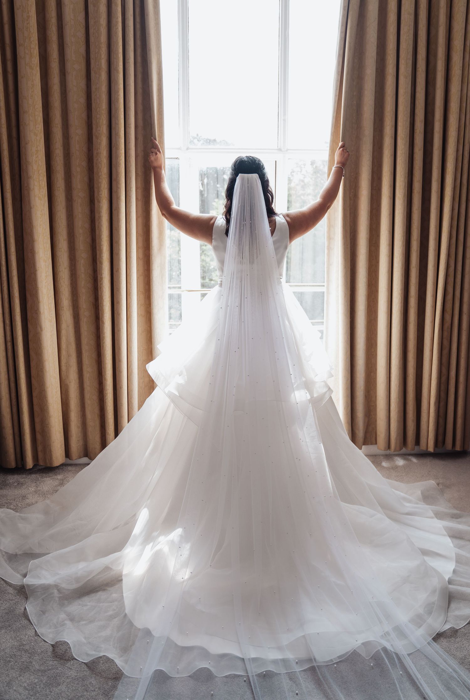 Back view of a bride in a flowing white wedding gown with long veil standing at a window.