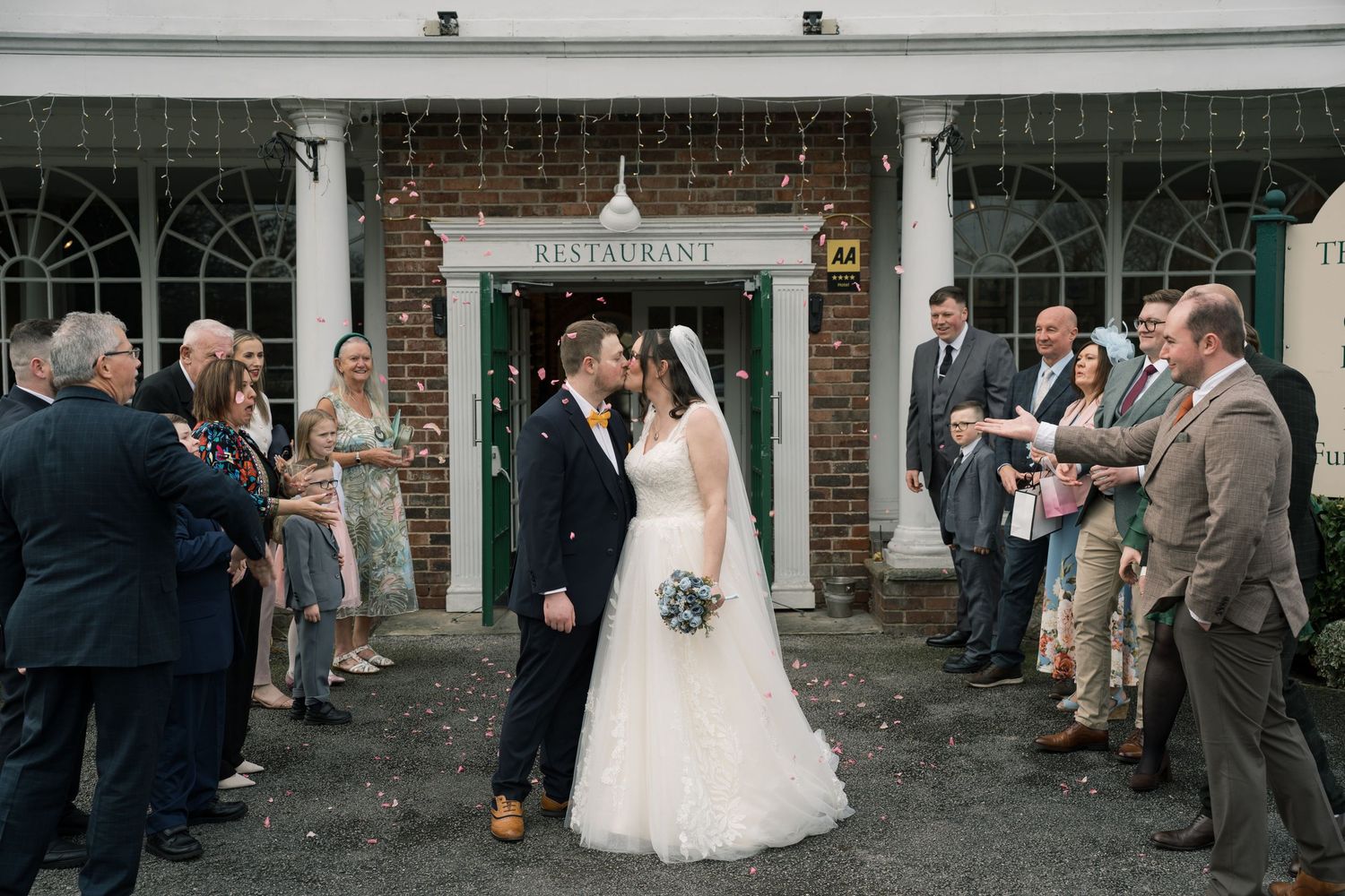 Wedding couple shares a kiss outside a brick building as guests line up to celebrate their special moment.