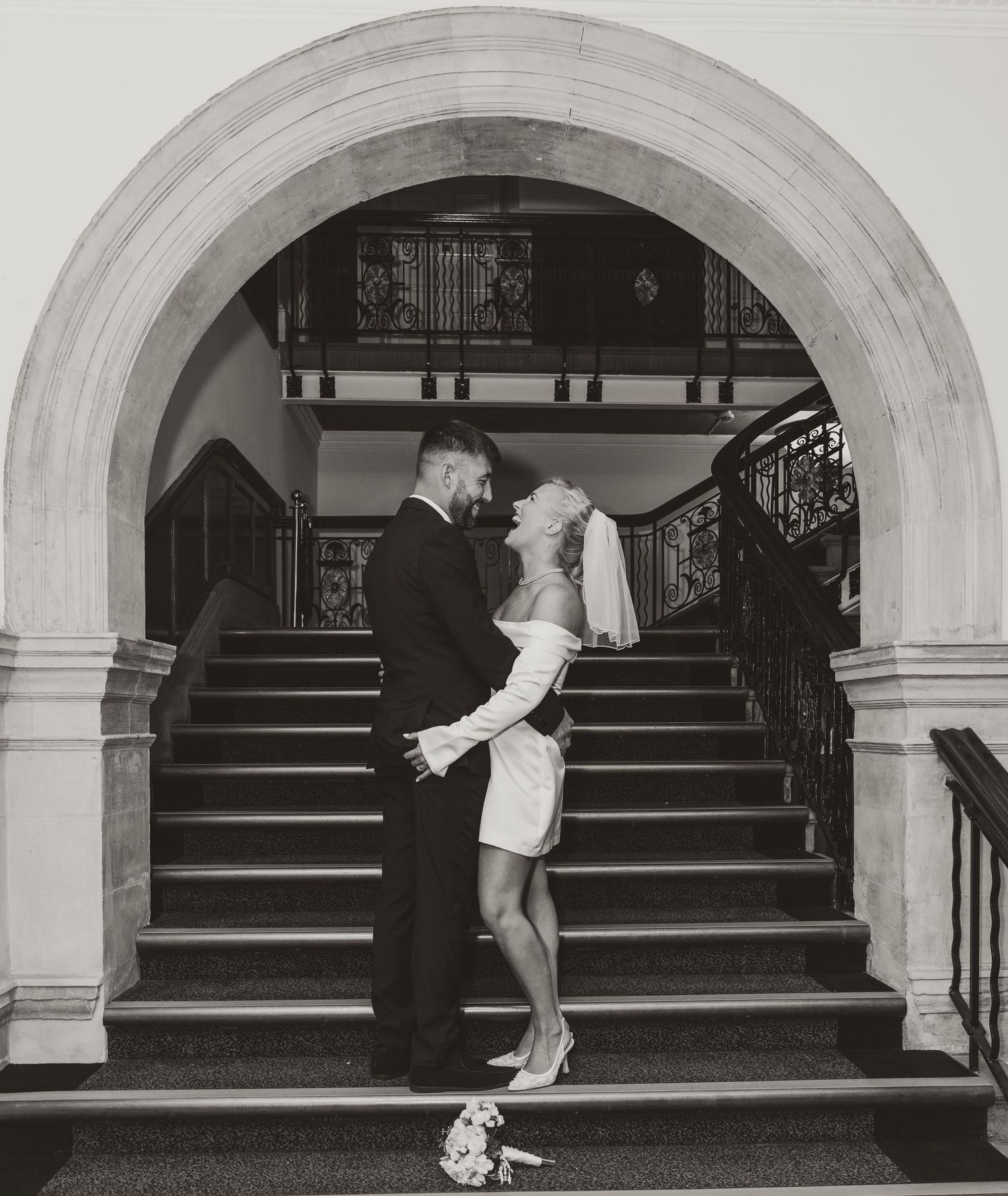 Black and white photo of a couple in wedding attire sharing a romantic moment under an arch by a grand staircase.