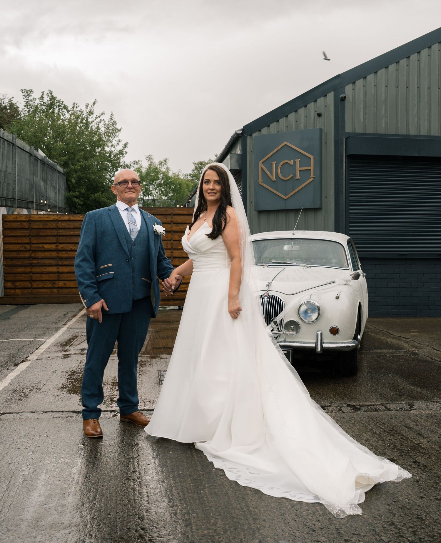 Bride in white gown poses with classic white vintage car outside industrial venue.