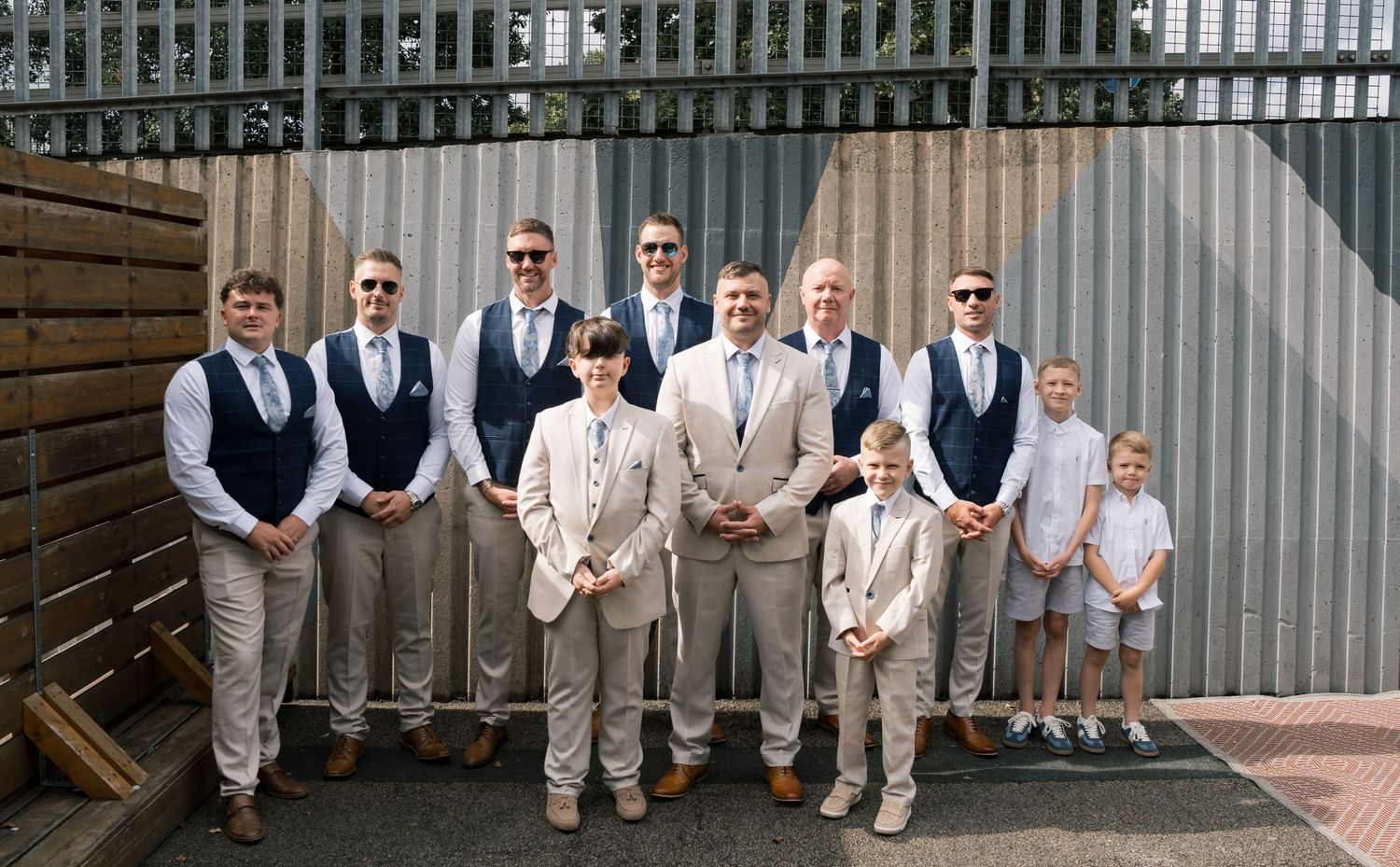 Wedding party members in coordinated khaki and navy attire pose casually against rustic backgrounds at outdoor venue.