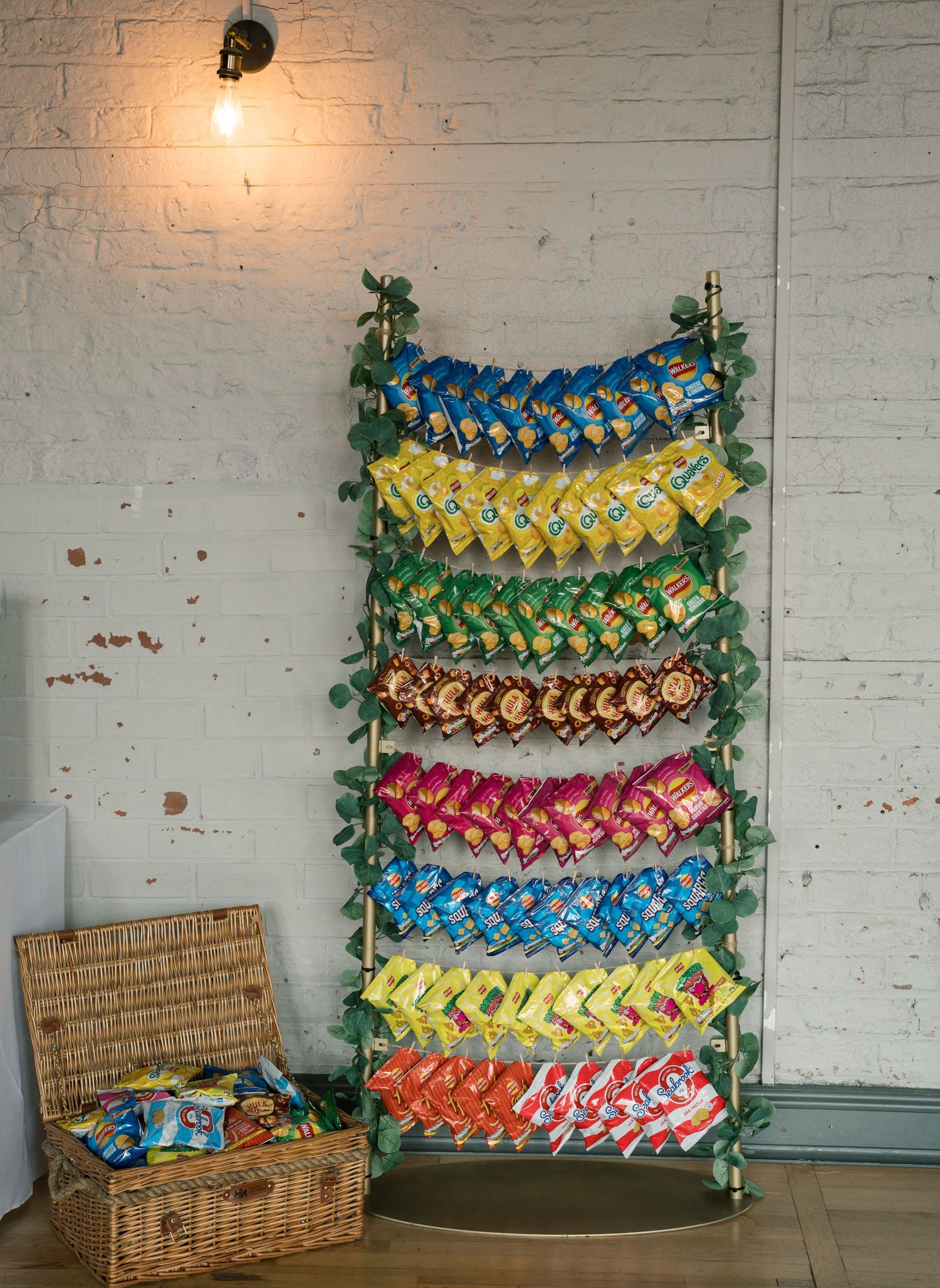 Colorful bags of snacks arranged in rainbow rows on a decorative ladder display against a white brick wall.