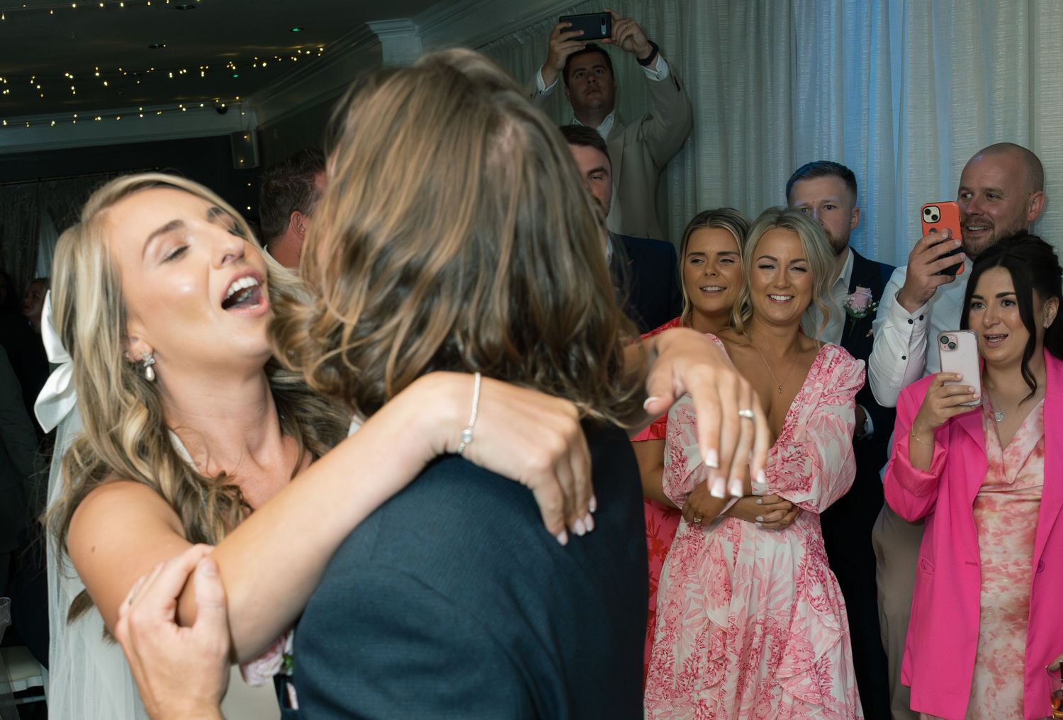 Wedding guests dance and celebrate under string lights at an indoor reception.
