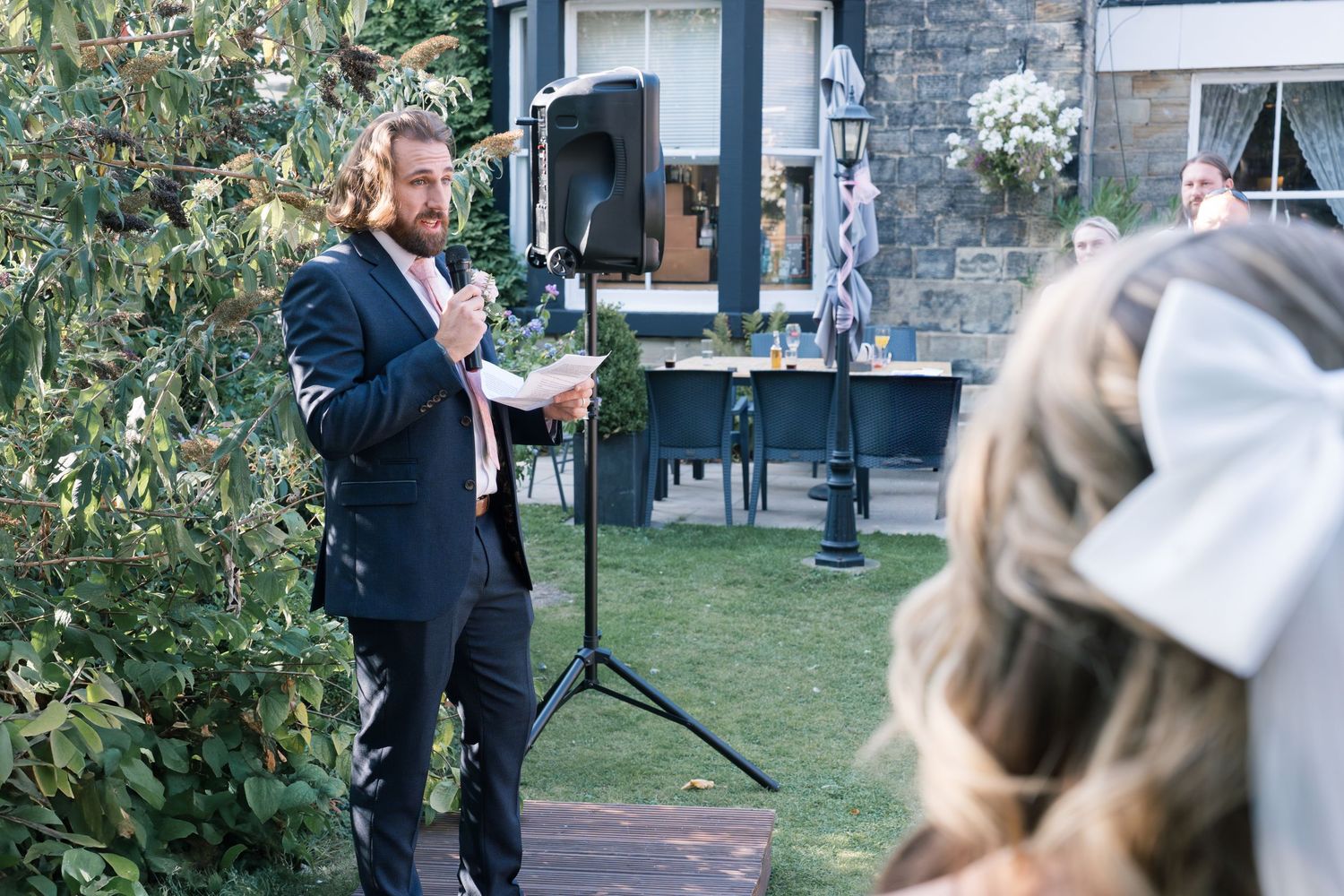Wedding ceremony in garden setting with speaker addressing guests outdoors.