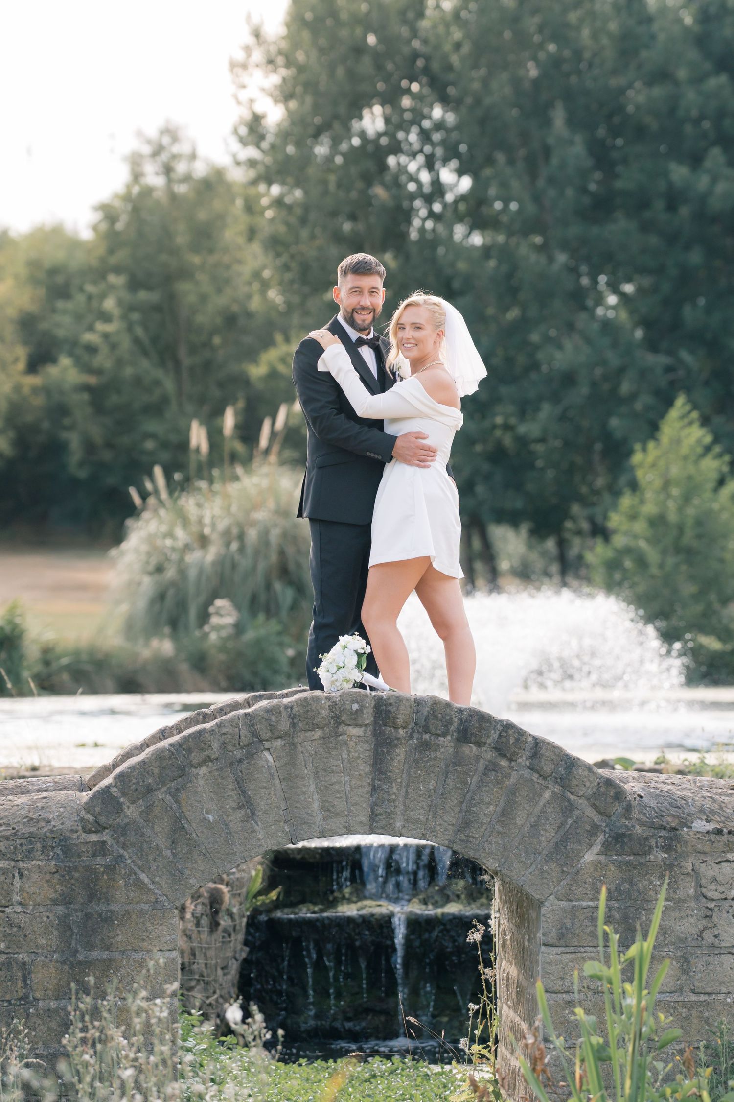 Bride and groom embracing on a stone arch bridge with a waterfall, surrounded by lush greenery on their wedding day.