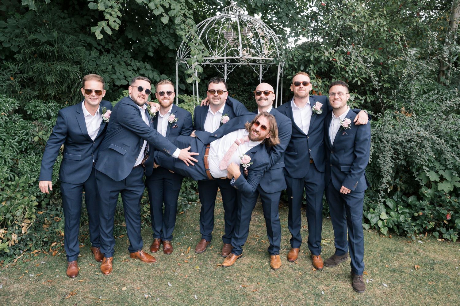 Groomsmen in navy suits and brown shoes posing together outdoors in a playful jumping shot.