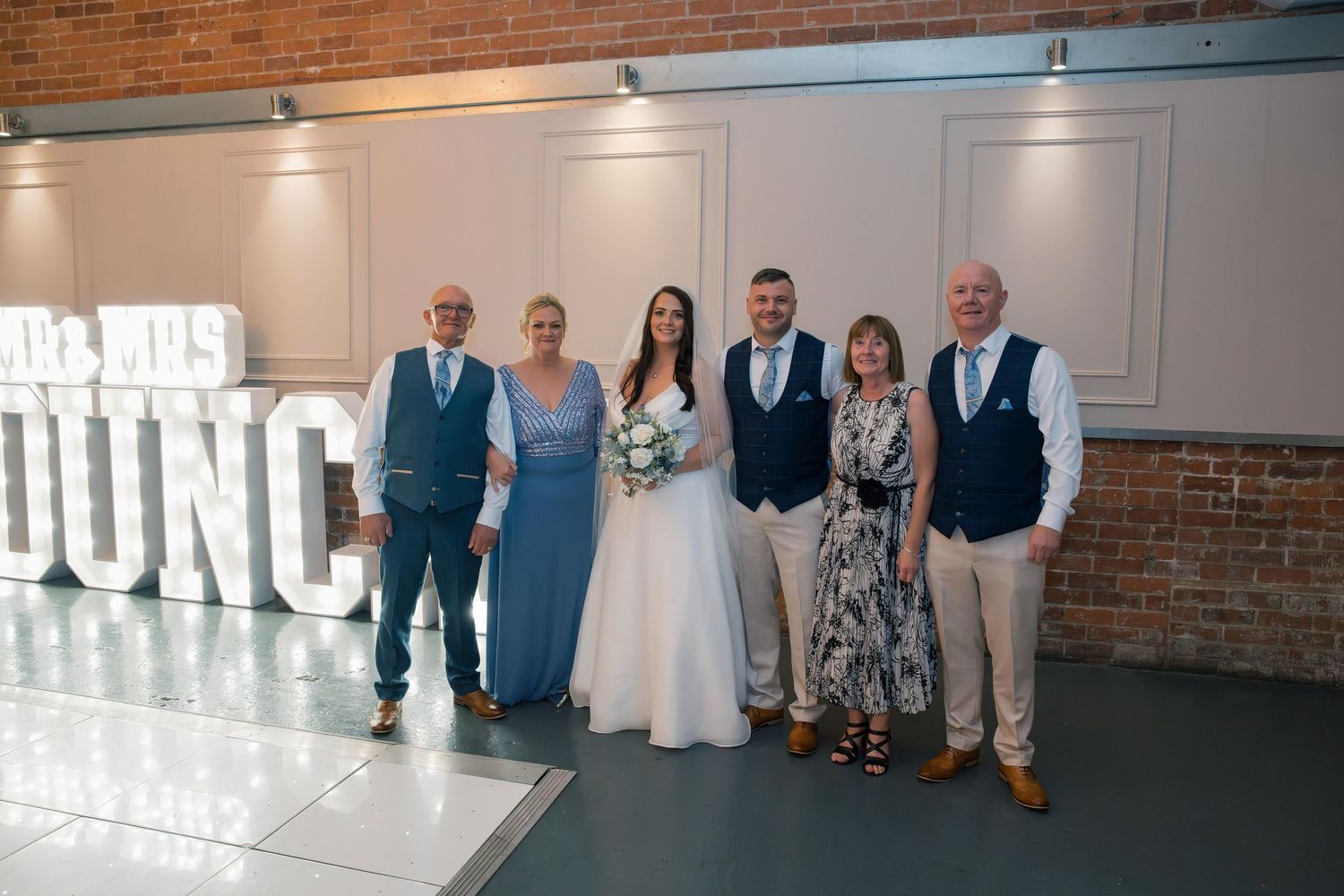 Wedding party lined up against white wall with large decorative letters in an industrial brick venue.