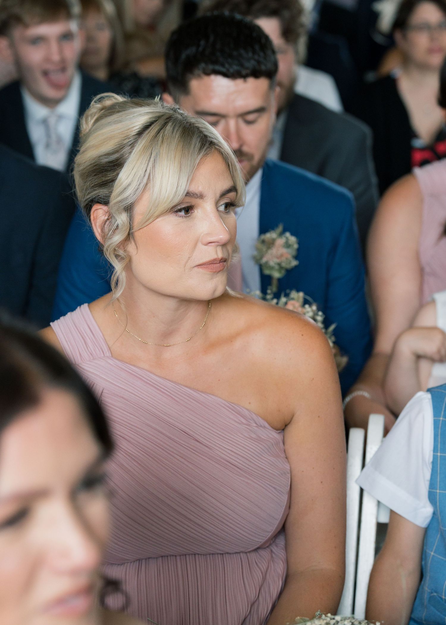 Bridesmaid in a mauve one-shoulder dress smiles during a wedding ceremony.