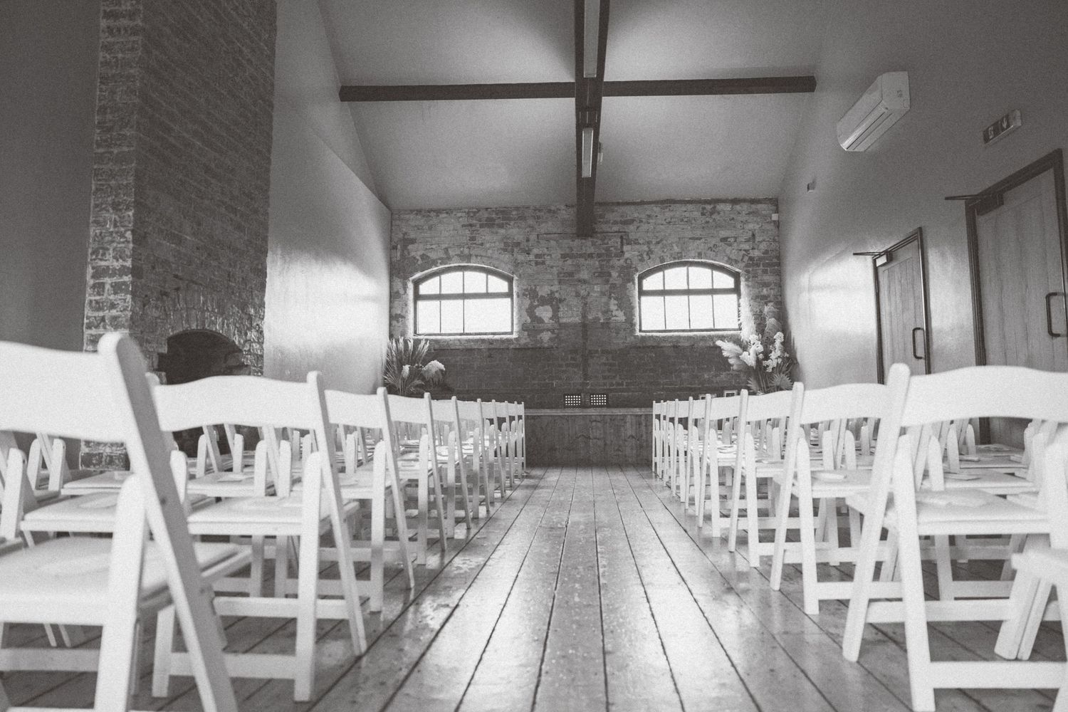 Black and white interior view of a church sanctuary with rows of white wooden chairs and exposed brick walls.