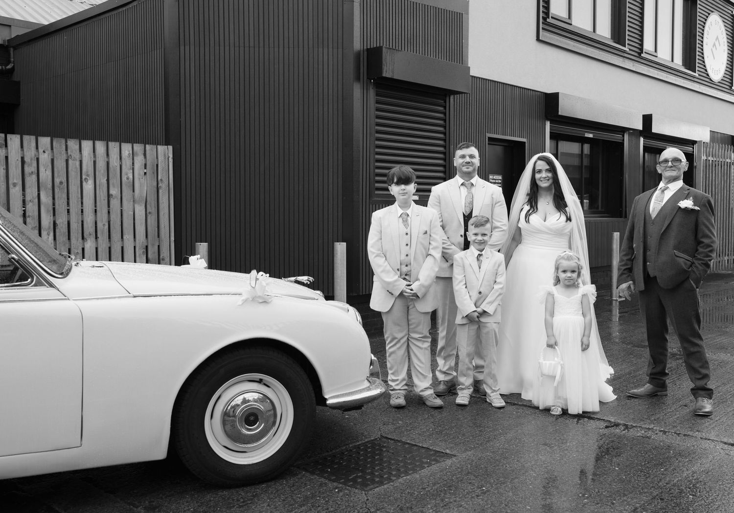Black and white panoramic shots of wedding party with vintage car outside industrial venue.