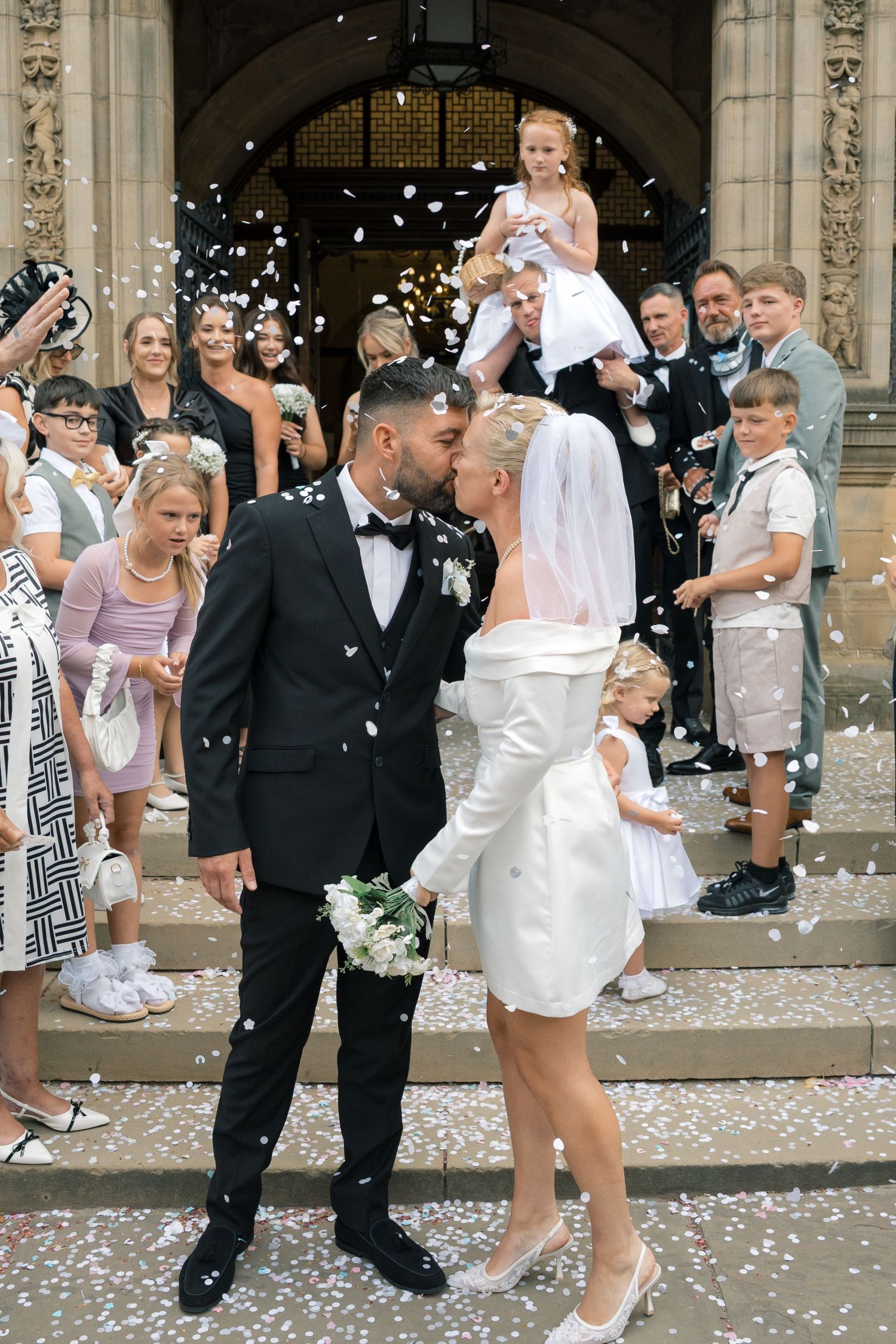 Newlywed couple kissing on church steps surrounded by guests throwing white confetti at their wedding.