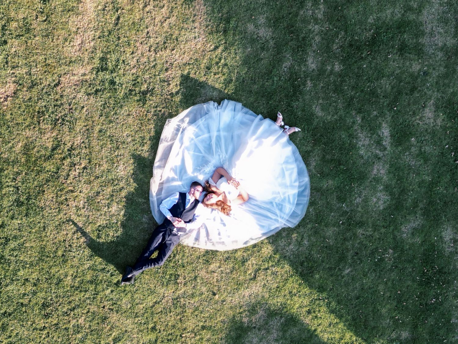 Aerial drone view of wedding couple lying on green grass, with bride in white dress forming a circular shape.